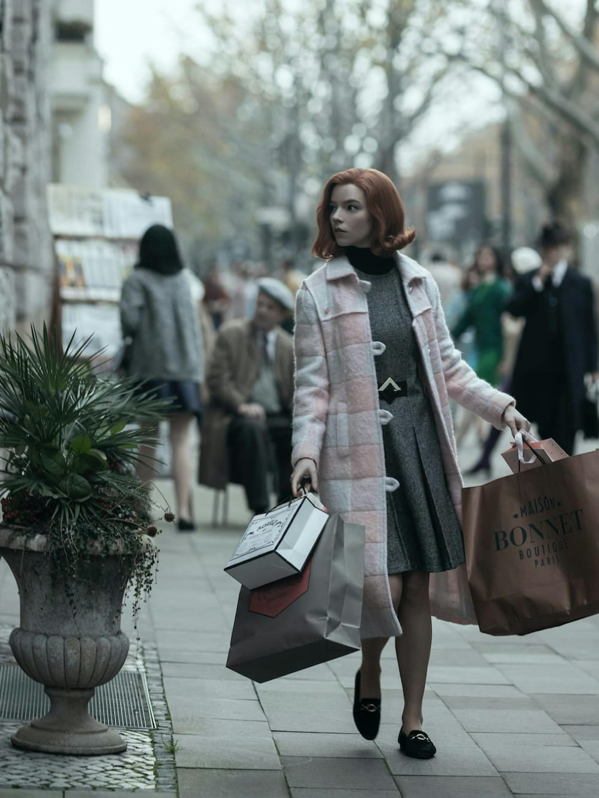 Beth Harmon (Anya Taylor-Joy) carries four shopping bags as she walks the streets of Paris. She wears a long white-and-pink checkered jacket that I’d love to borrow.