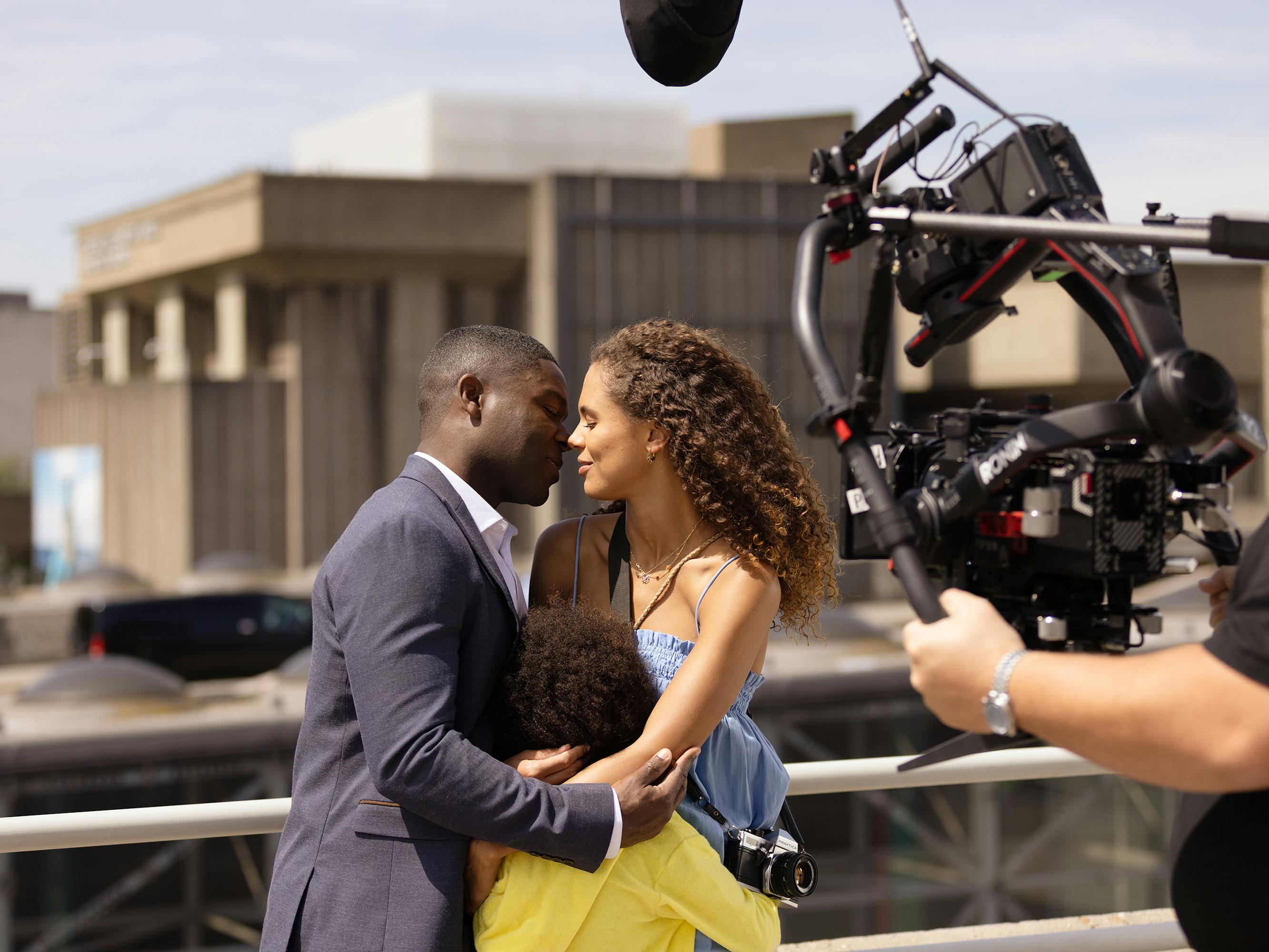 Dayo (David Oyelowo), Laura (Amelie Dokubo), and Amanda (Jessica Plummer) stand together on a bridge. David wears a navy suit, Amanda wears a strappy blue dress, and Laura wears a neon yellow hoodie.