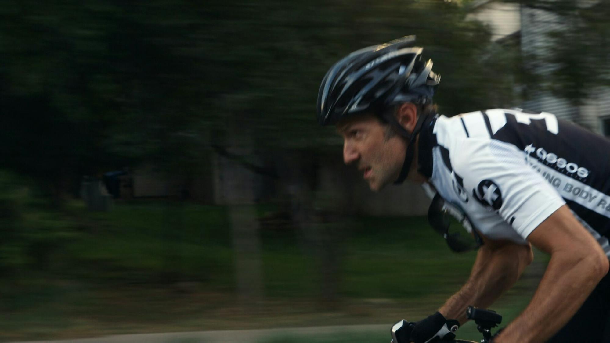Bryan Fogel speeds by greenery on a bike. His shirt is black and white and his helmet is also black. He looks intense!