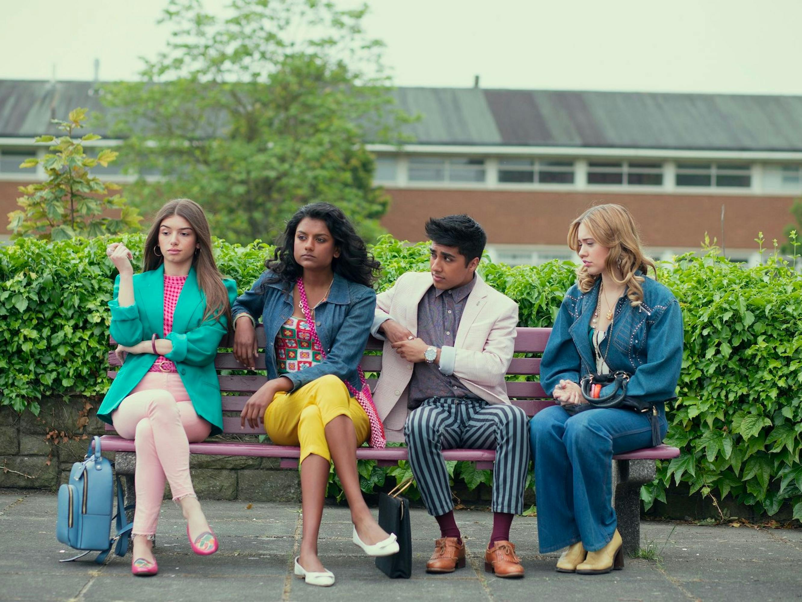 Ruby Matthews (Mimi Keene), Olivia Hanan (Simone Ashley), Anwar (Chaneil Kular), and Aimee Gibbs (Aimme Lou Wood) sit on a red bench together looking fabulous. Behind the bench is a row of green hedges and in the backdrop is Moordale Secondary School.