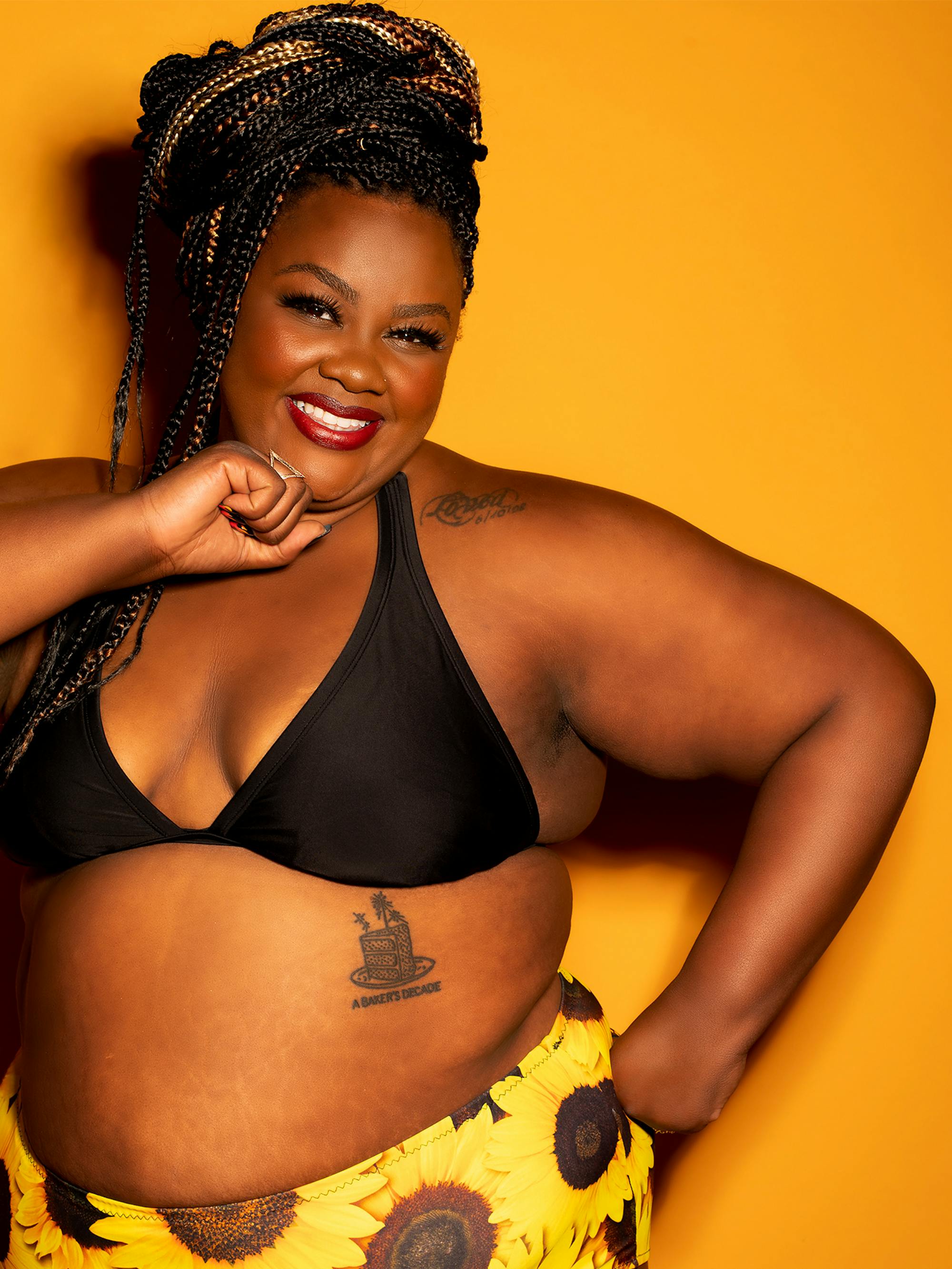 Nicole Byer poses in front of an orange yellow wall, wearing a bikini with sunflower print, smiling directly at the camera.