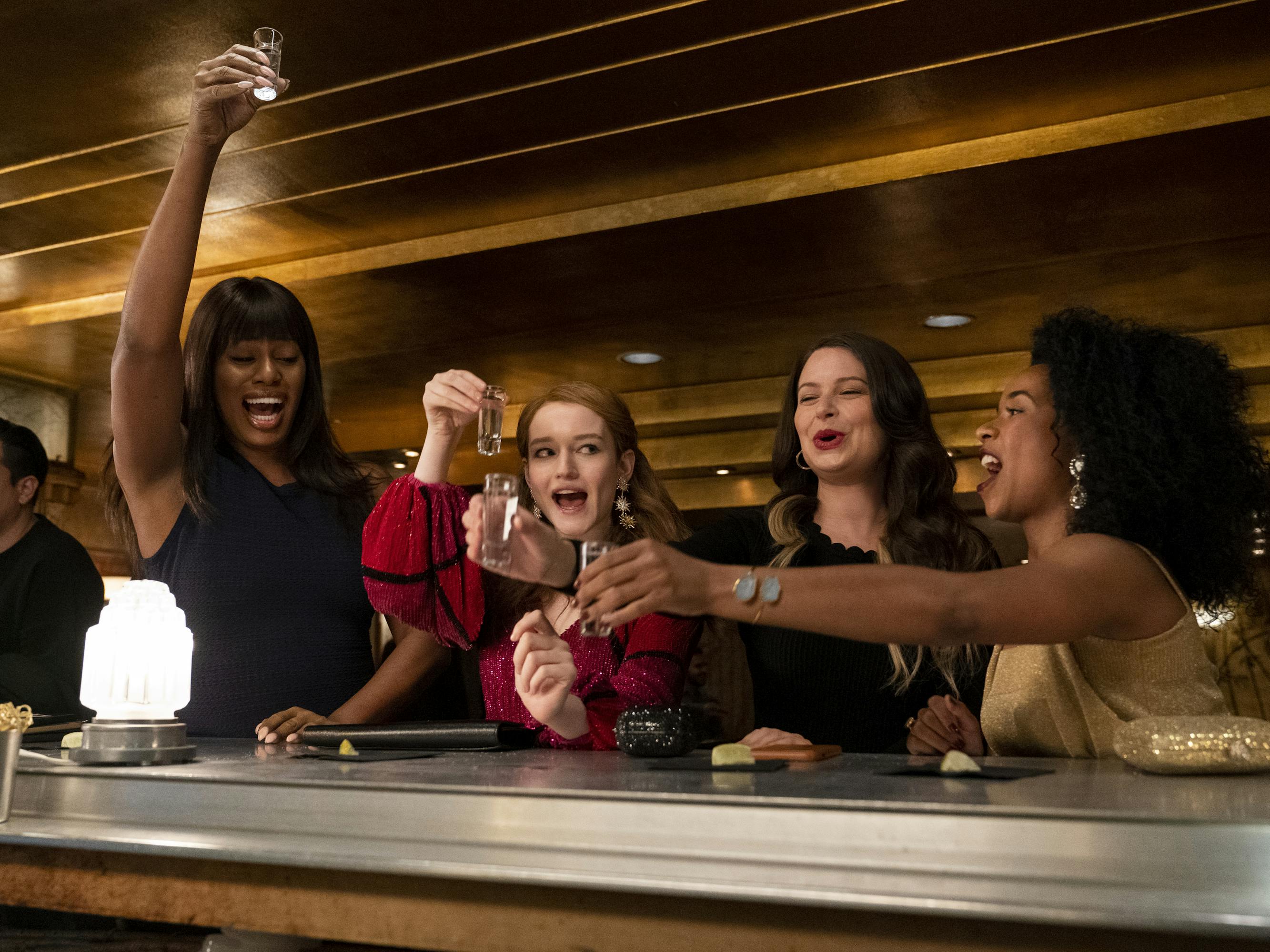 Kacy (Laverne Cox), Anna Delvey (Julia Garner), Rachel (Katie Lowes), and Neff (Alexis Floyd) take shots at a dimly lit bar. Cox wears a black dress, Garner wears red, Lowes wears a black gauzy top, and Floyd wears a cream top with matching clutch.