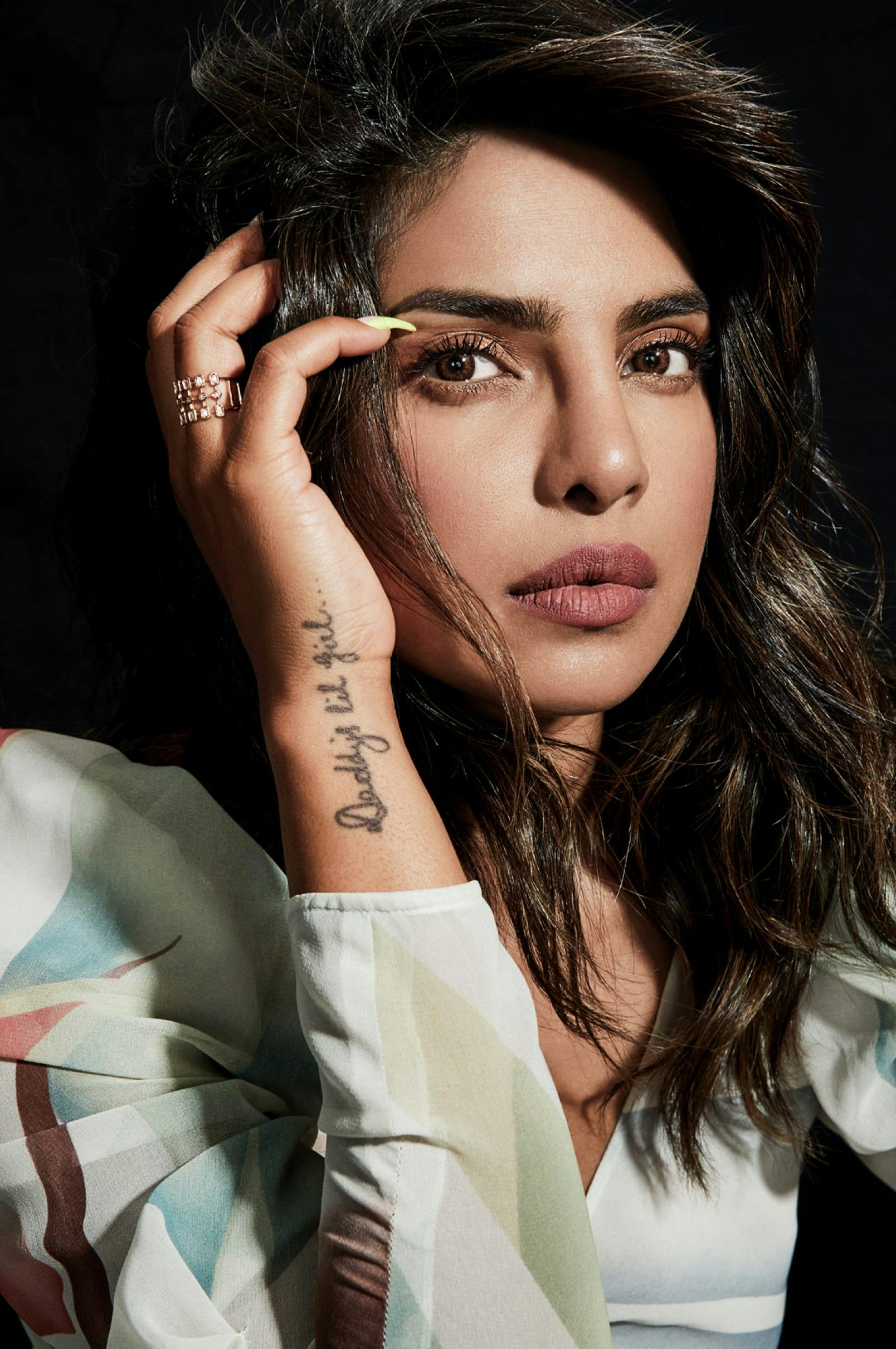 A portrait of Priyanka Chopra Jonas against a black backdrop. She wears a colorful dress with puff sleeves, and a tattoo is visible on her forearm as she runs her hand through her hair. 