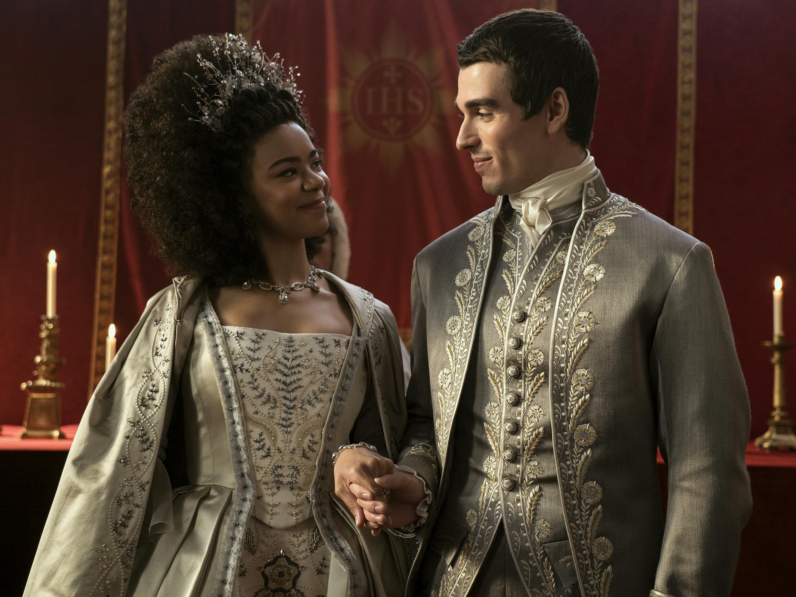 Queen Charlotte (India Amarteifio) and King George (Corey Mylchreest) wear silver ensembles and stand in a red draped room lit by taper candles.