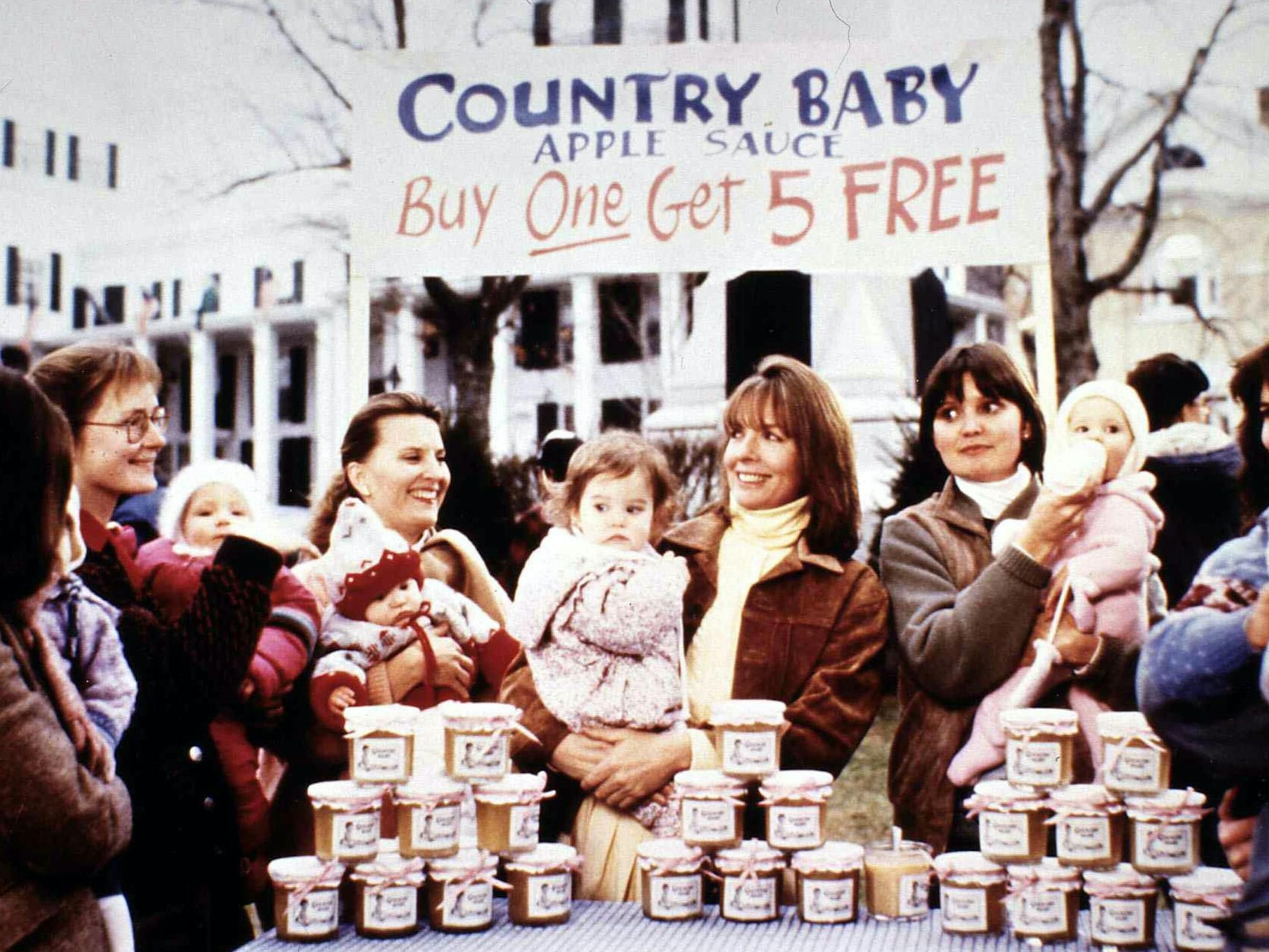 J.C. Wiatt (Diane Keaton) and the cast of Baby Boom. She holds her baby behind a table with piles of applesauce. In the background is a banner that reads: Country Baby Apple Sauce Buy One Get 5 Free.