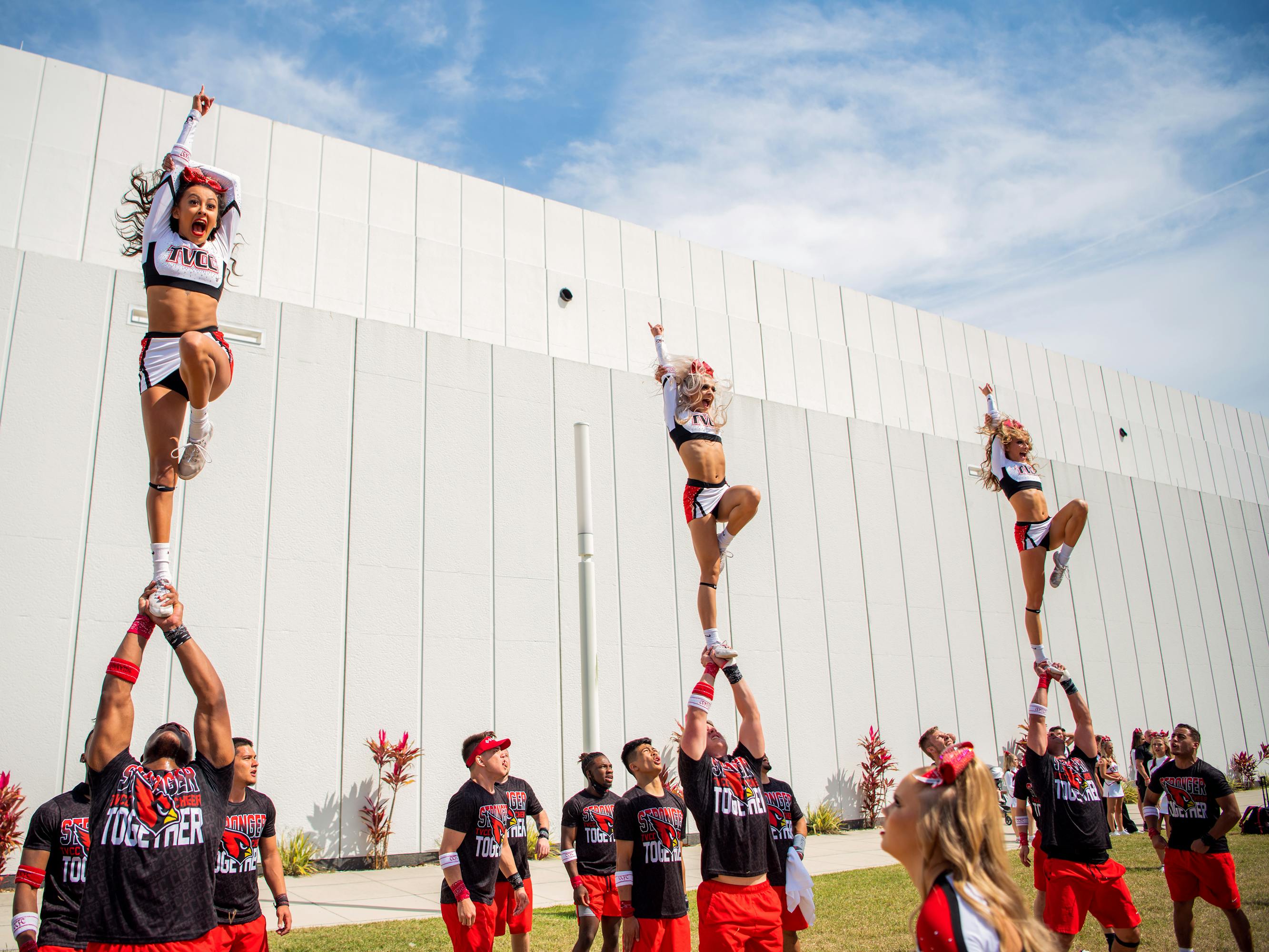 Trinity Valley Community College Cheer team performs an impressive stunt. Men in red shorts and black shirts stand around, and three hold women above their heads on one single foot. This looks so hard to balance!