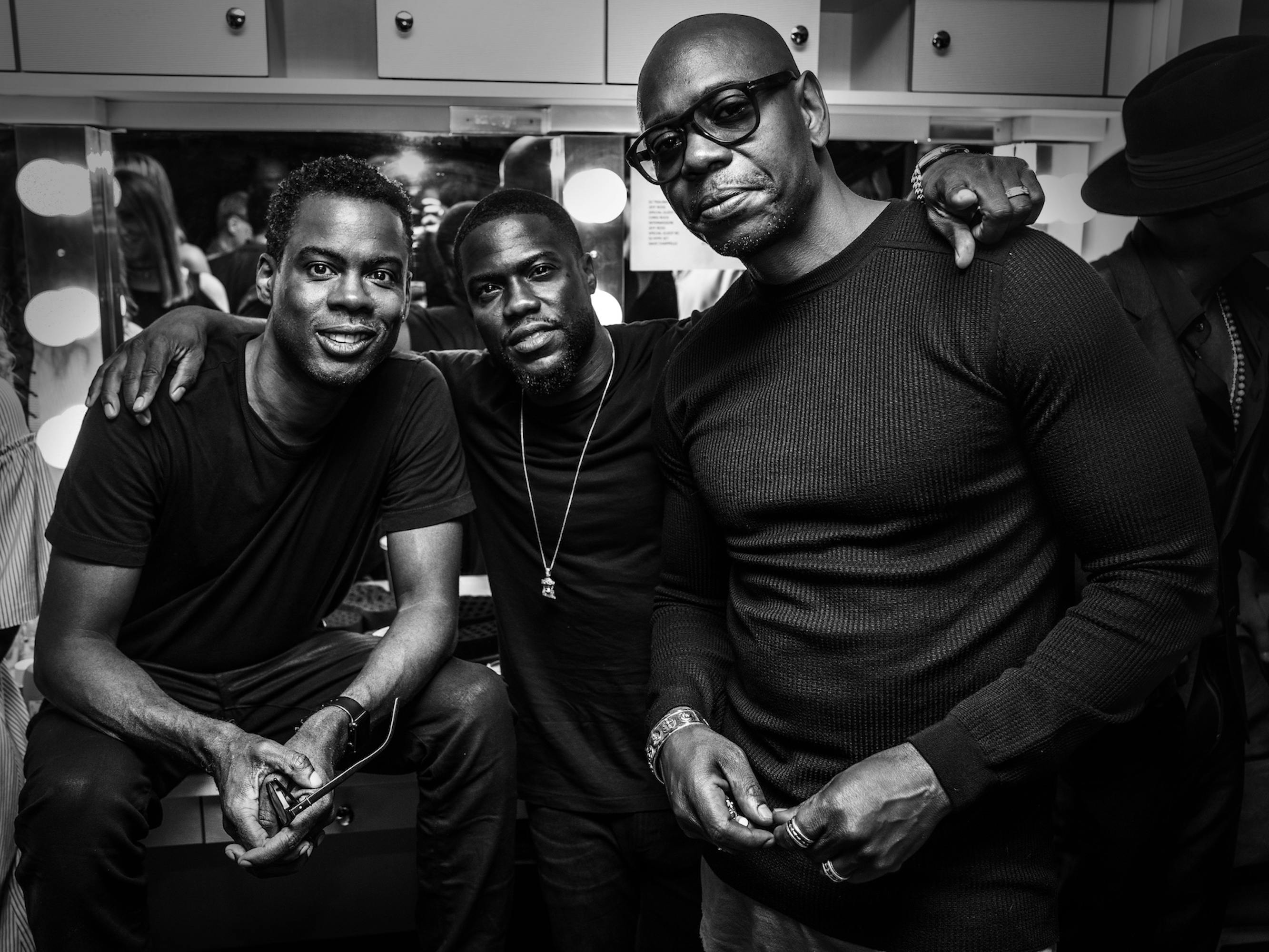 Chris Rock, Kevin Hart, and Chappelle stand in all black outfits, with their arms around each other.