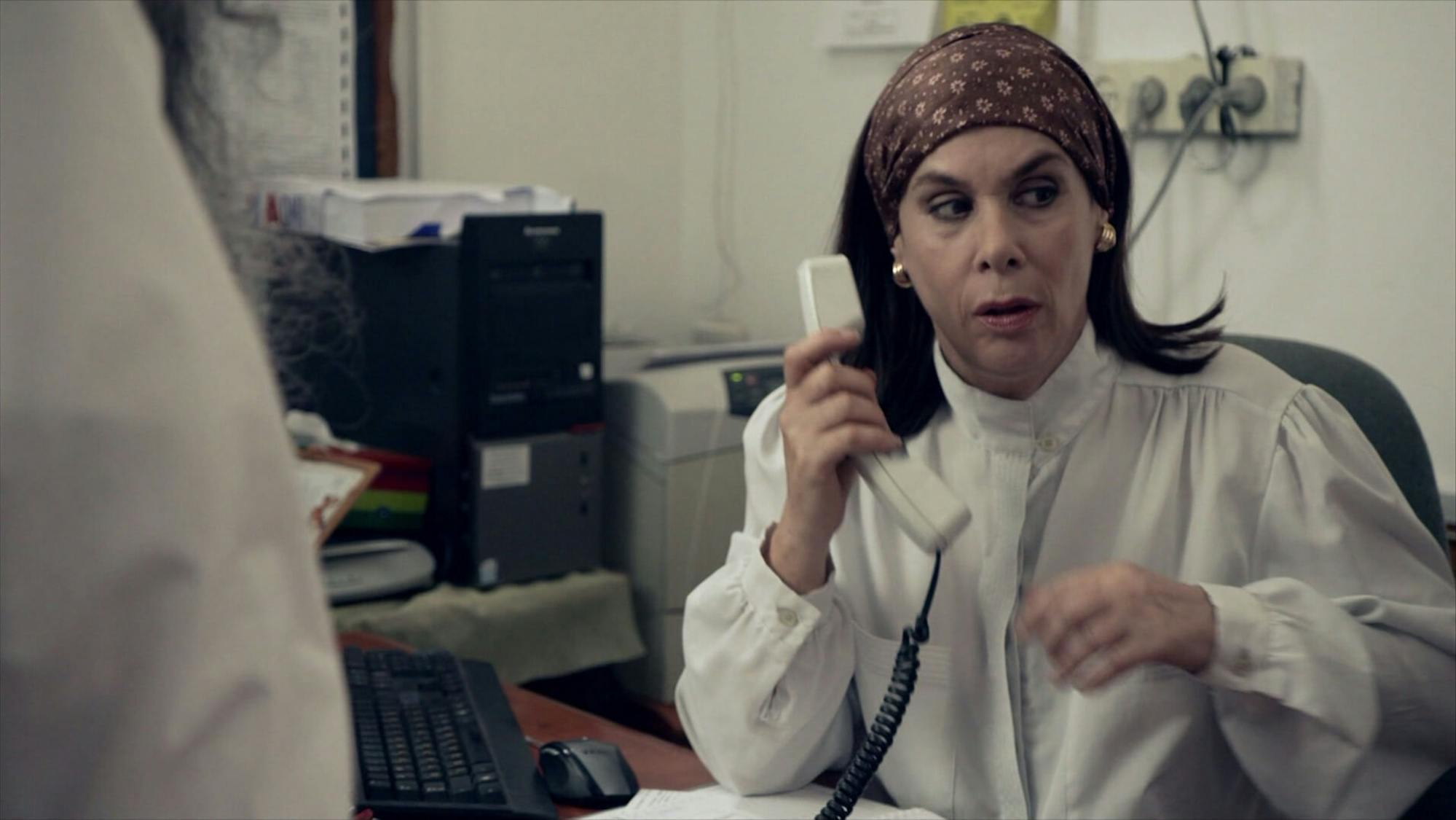 Aliza Gvili (Orly Silbersatz) wears a white blouse, gold earrings, and a silk headband. Her hair is curled at the ends, and she manages a desk with computers, files, printers, and keyboards. On the left side there is a figure’s arm, making the scene seem ominous.