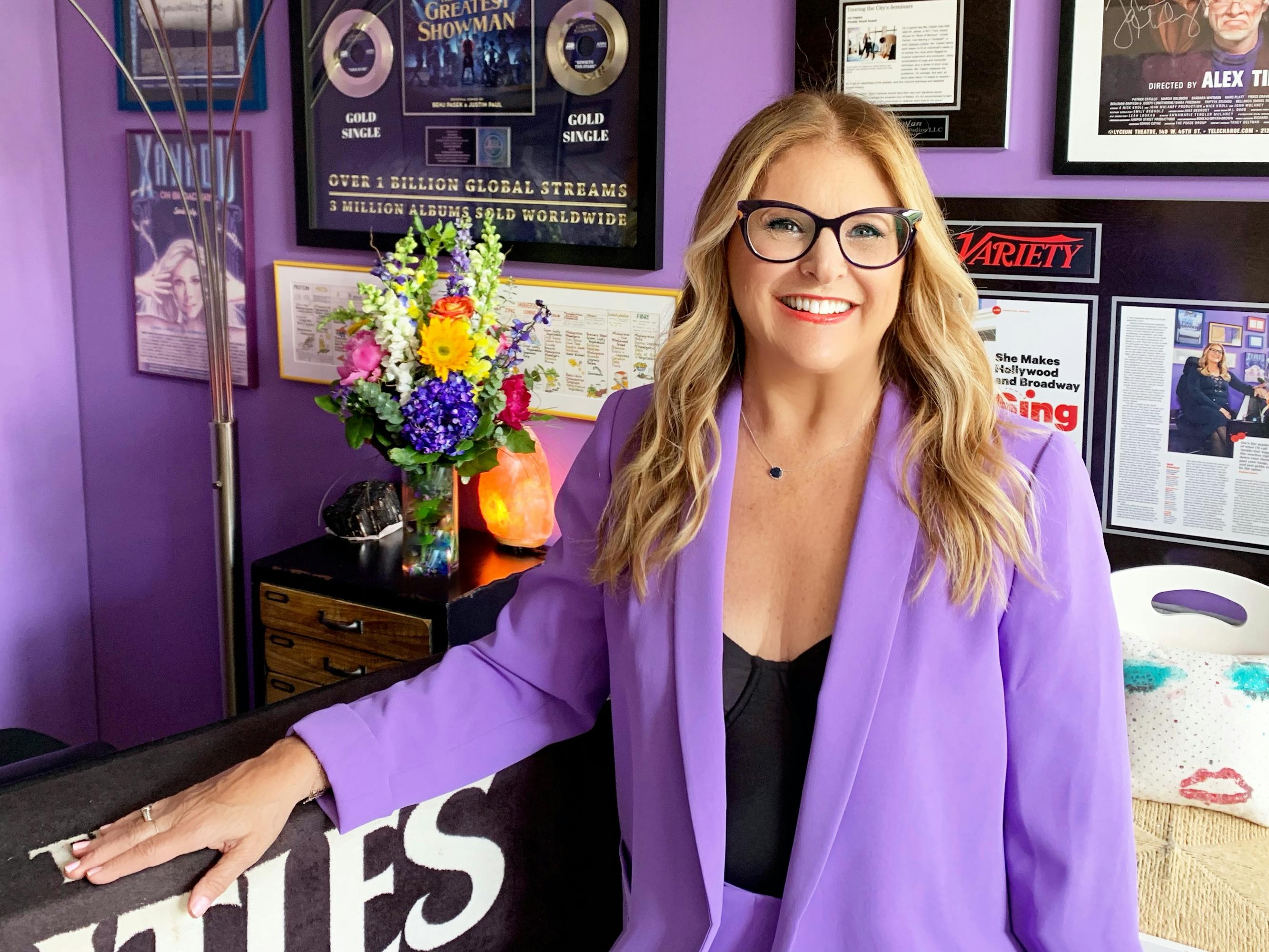 Liz Caplan stands in front of awards plaques reading "Greatest Showman, Over 1 billion Global Streams, 3 million albums sold worldwide" and "She Makes Hollywood and Broadway." Caplan is wearing a lavender blazer and black glasses.