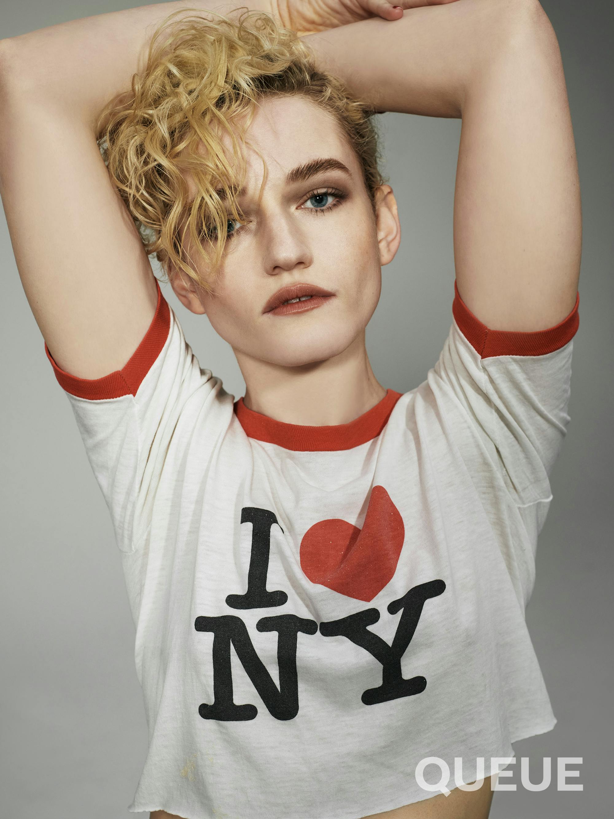 Julia Garner wears a I Love New York shirt and rests her arms above her head.