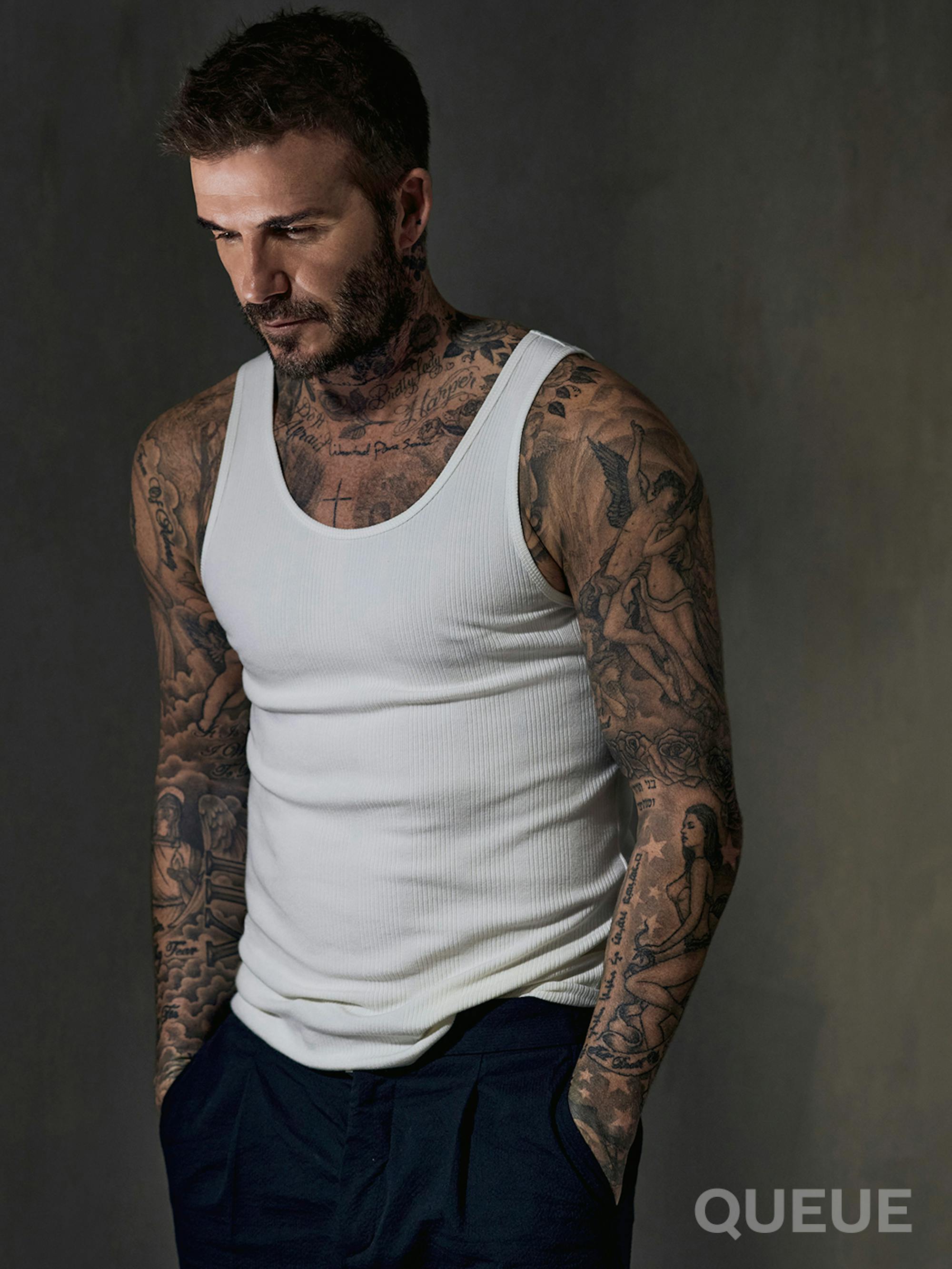 David Beckham wears black pants and a white shirt in a gray-walled room.