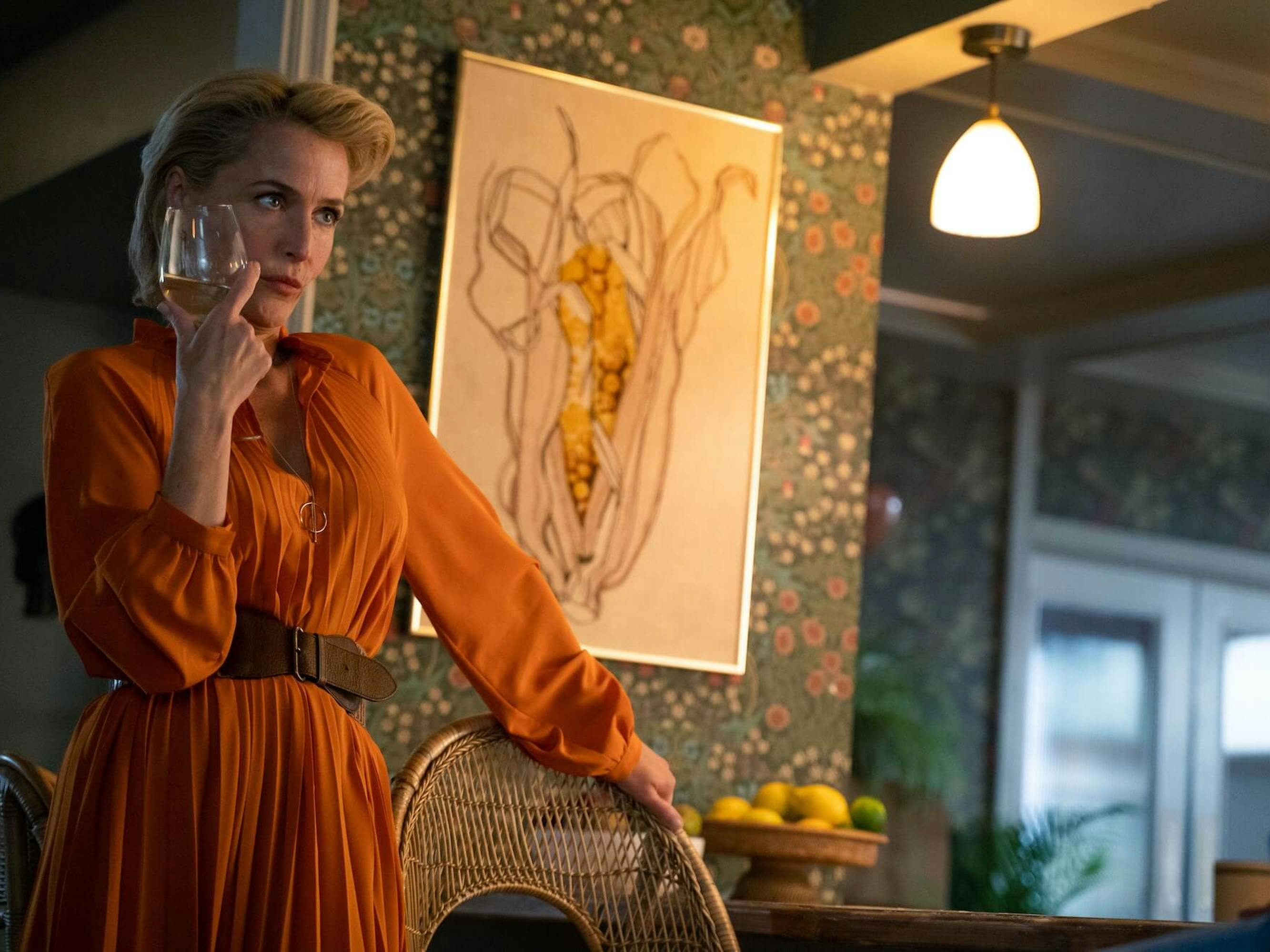 Jean Milburn (Gillian Anderson) in Sex Education stands next to a wicker chair wearing an orange dress and holding a glass of white wine.