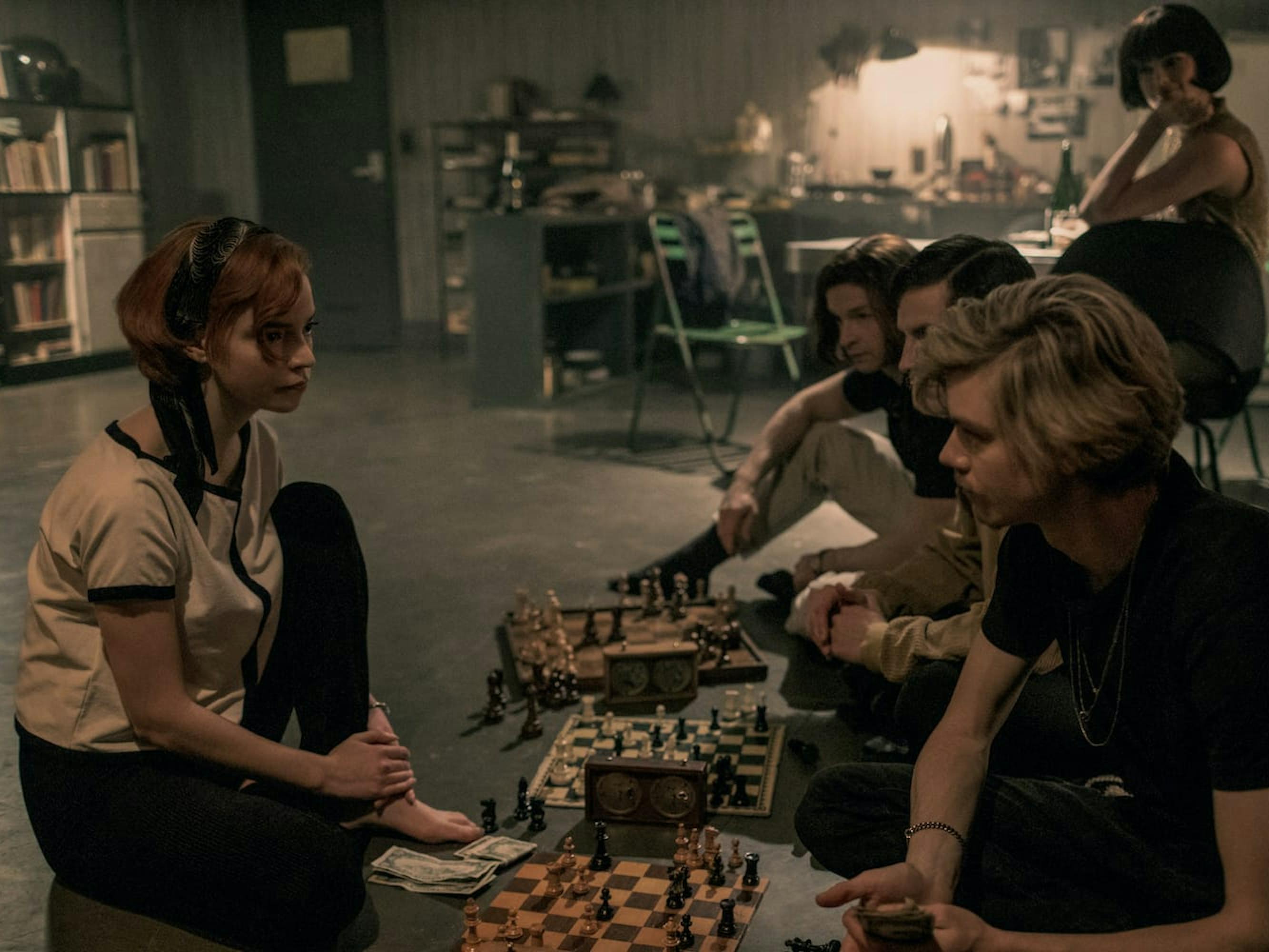 Beth Harmon (Anya Taylor-Joy) and Benny Watts (Thomas Brodie-Sangster) sit ina basement and play chess as three others look on. There are three chess boards on the ground, and green chairs scattered in the background.
