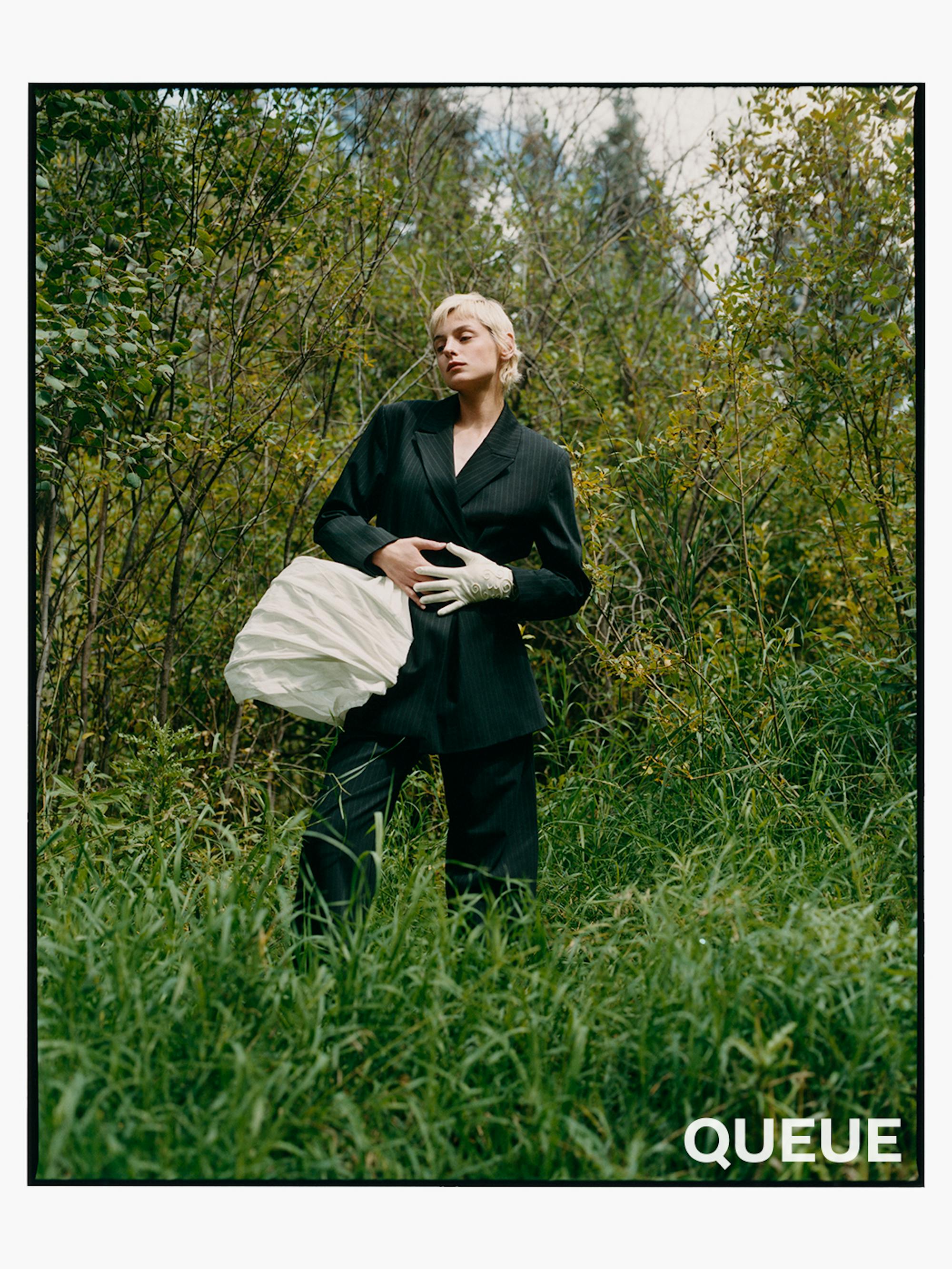 Emma Corrin wears a black ensemble with a white side piece, standing in a green field.