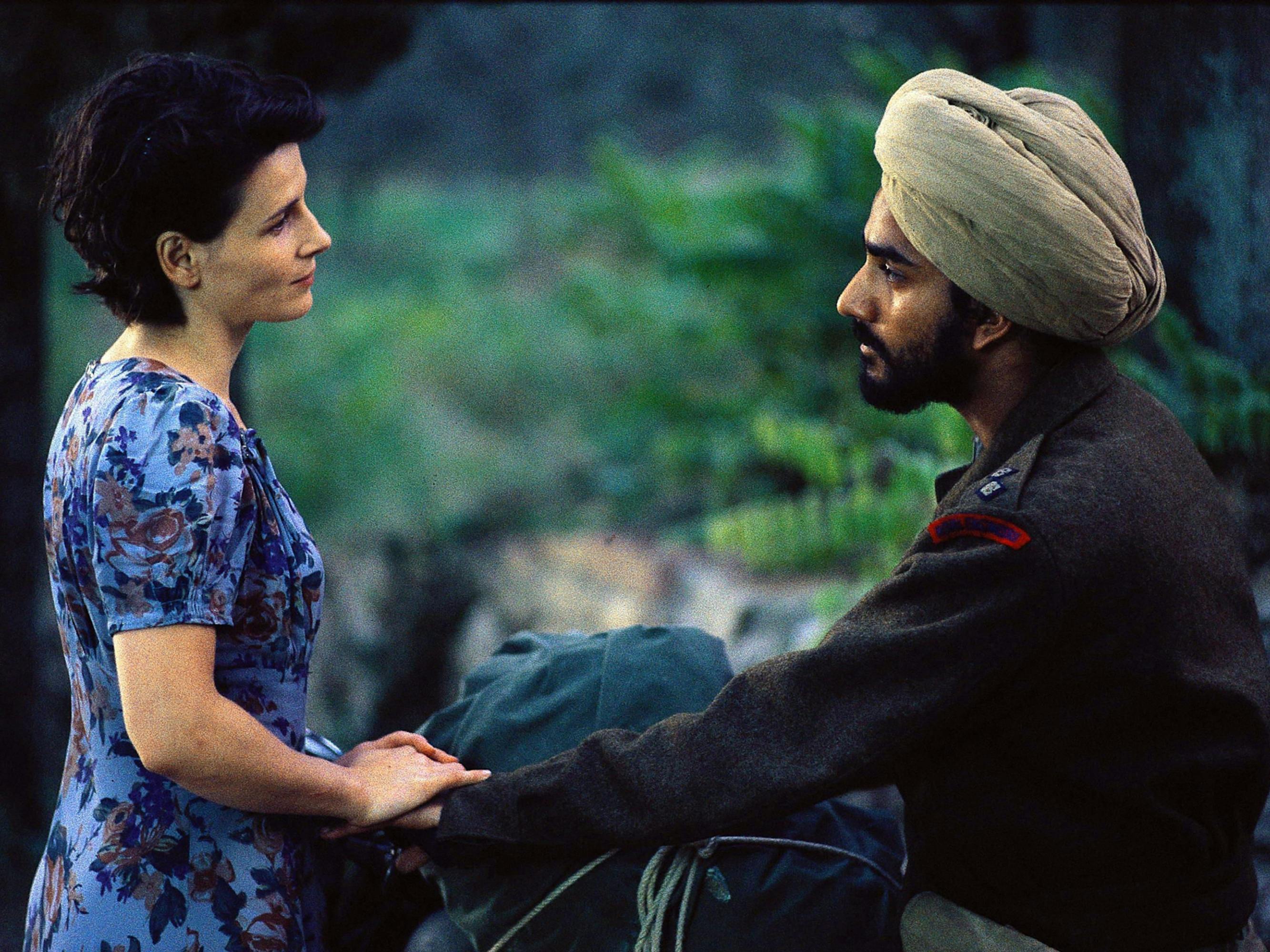 Juliette Bincohe holds the hand of Naveen Andrews in The English Patient. She wears a blue floral dress; he is in a dark jacket with red trimmed lapels and wears a beige turban. They look at each other fondly. 