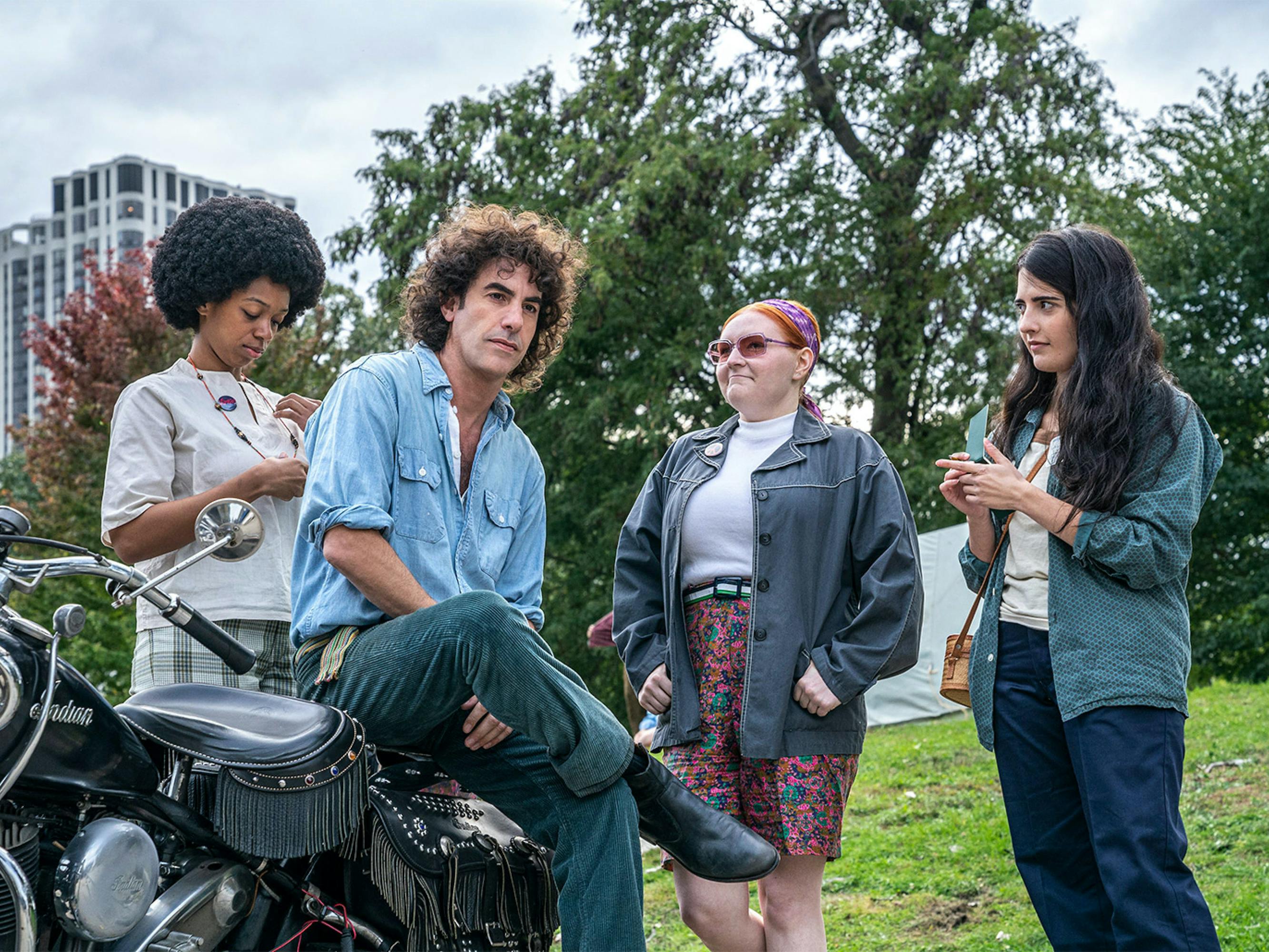 The actor sits on a motorcycle, surrounded by three other actors in retro 60s clothing. He wears a jean shirt, black leather boots, and his hair looks wild.