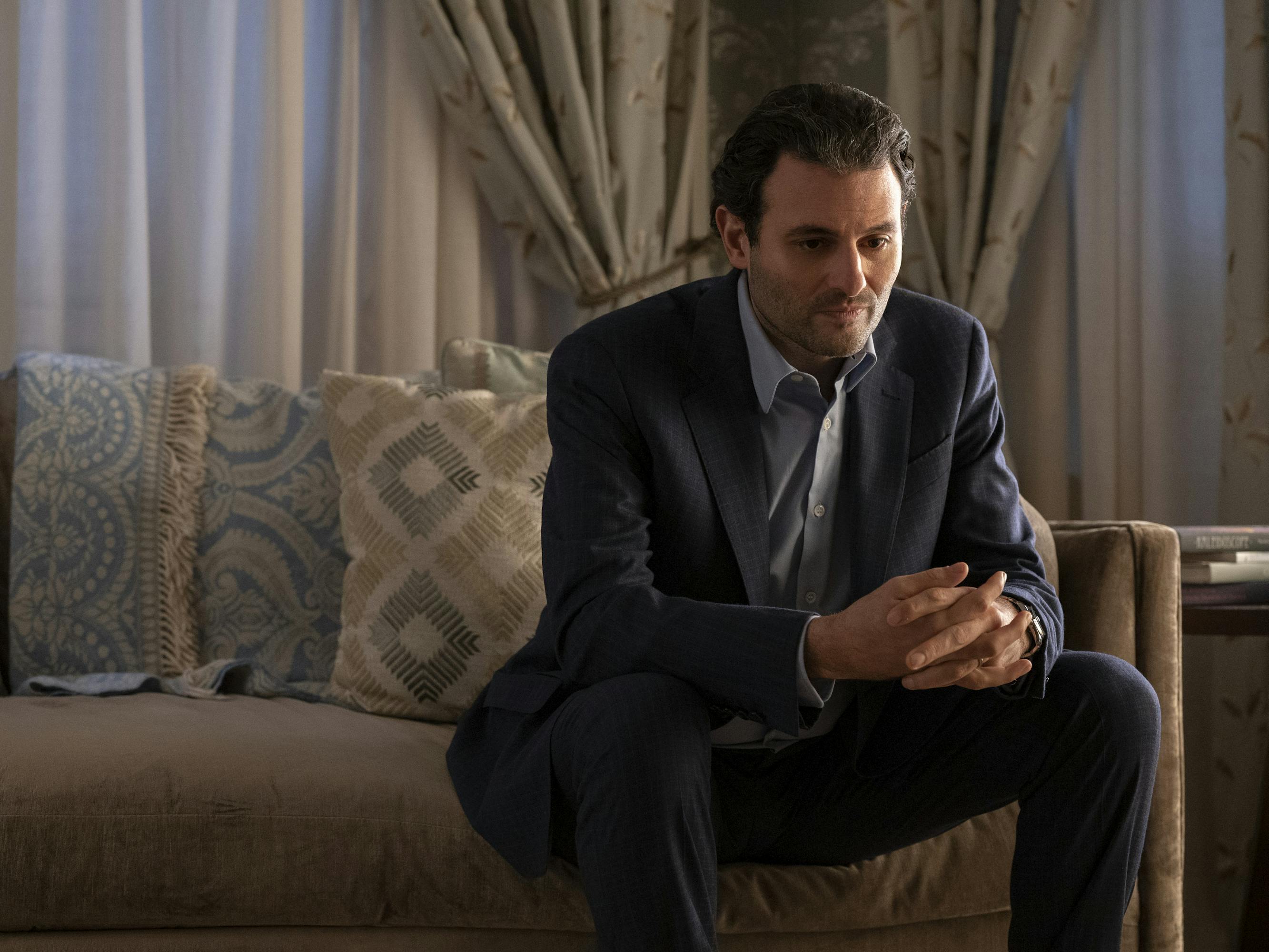 Arian Moayed wears a dark suit and sits on a beige couch with blue and beige pillows.