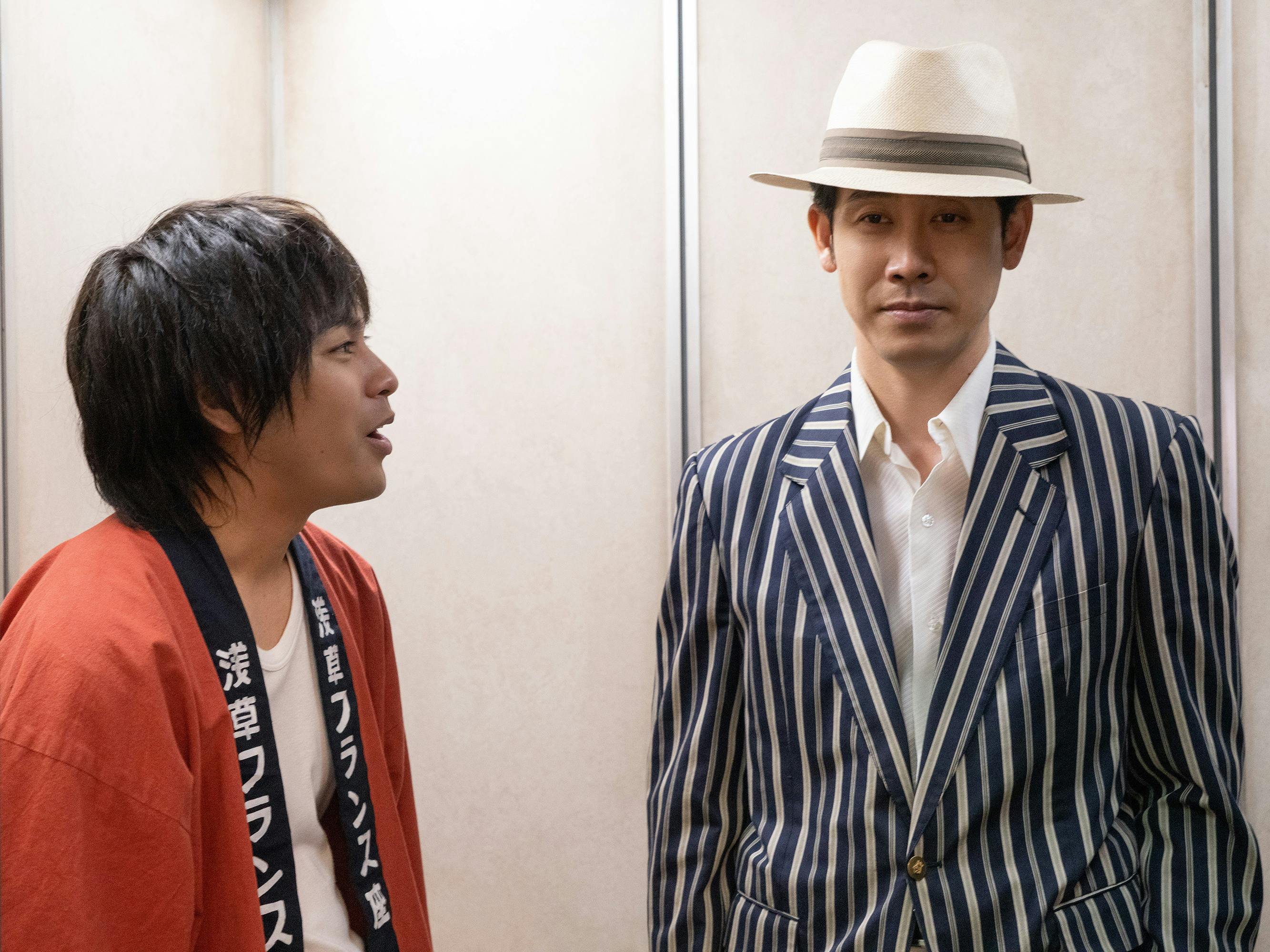 Yuya Yagira and Yo Oizumi stand in an elevator. Yagira wears a red robe and white shirt. Oizumi wears a white shirt, white hat, and black-and-white striped blazer.