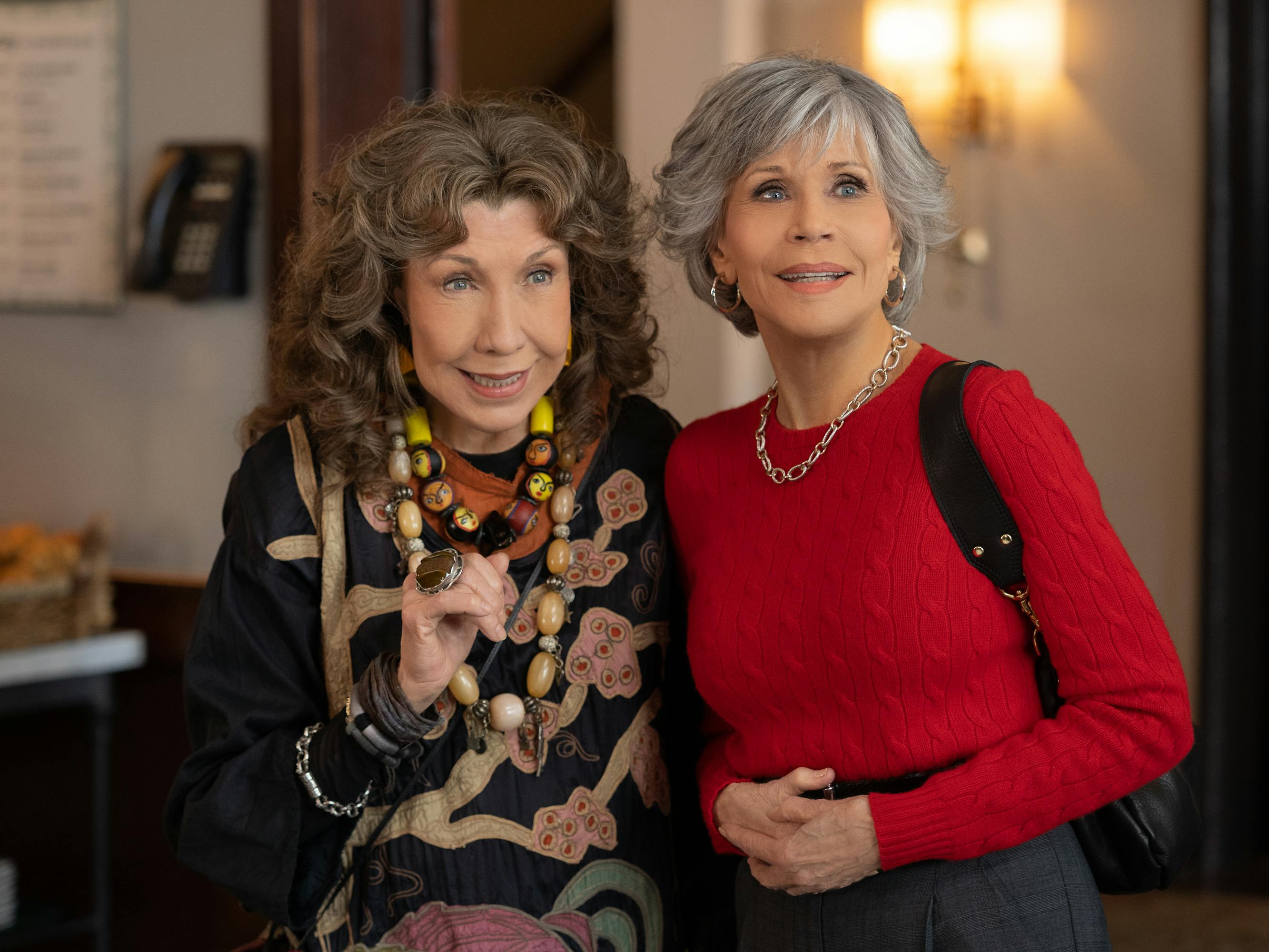 Frankie (Lily Tomlin) and Grace (Jane Fonda) stand close together. Frankie wears her assortment of chunky jewelry. And Grace wears a red sweater.