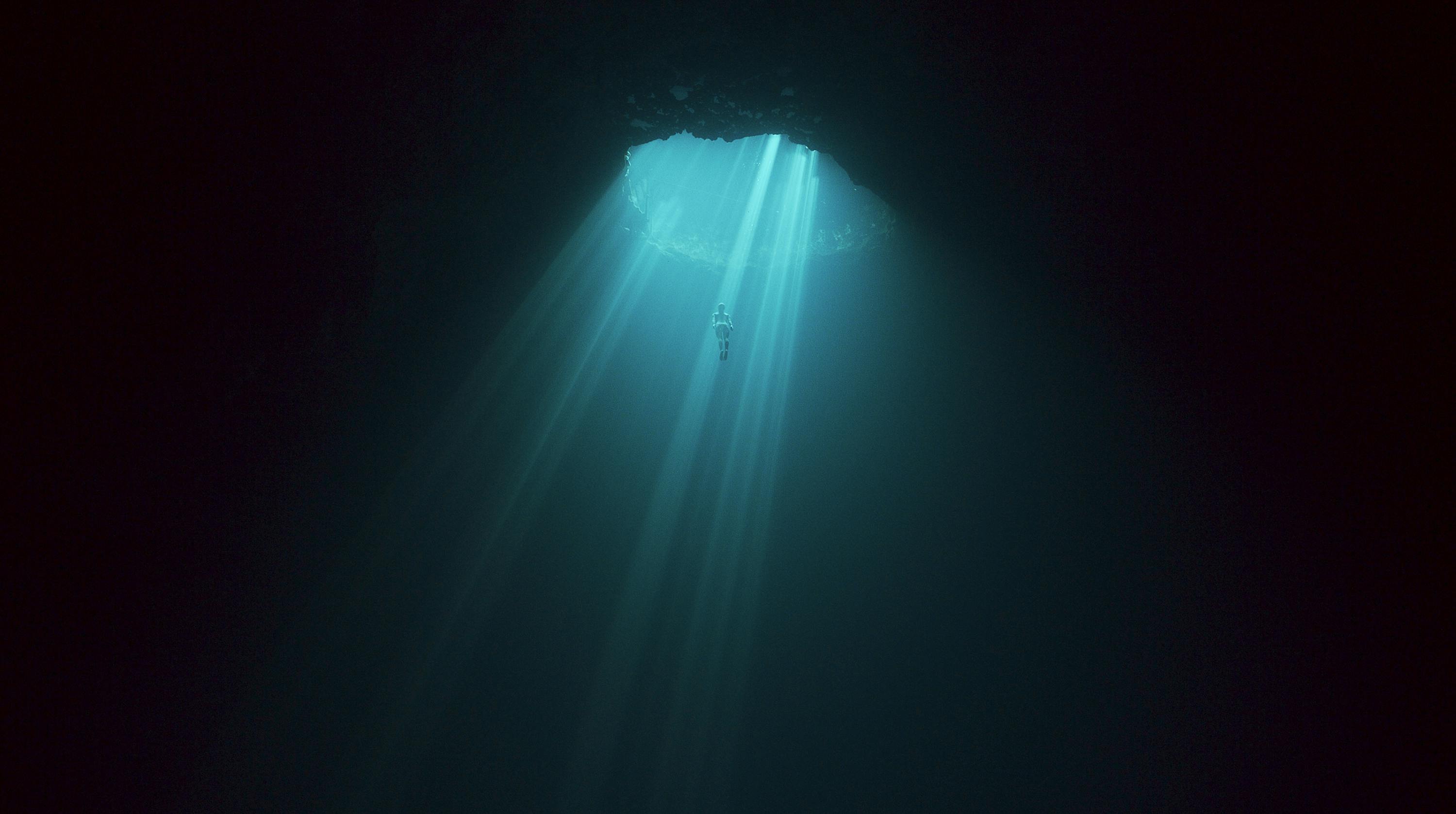 A diver appears lit by turquoise shafts of light in a darkened underwater shot.