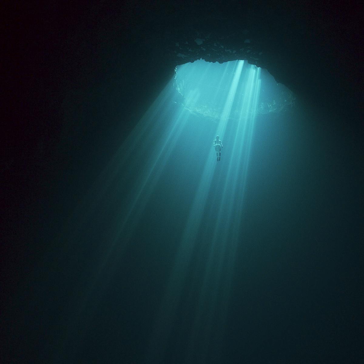 A diver appears lit by turquoise shafts of light in a darkened underwater shot.