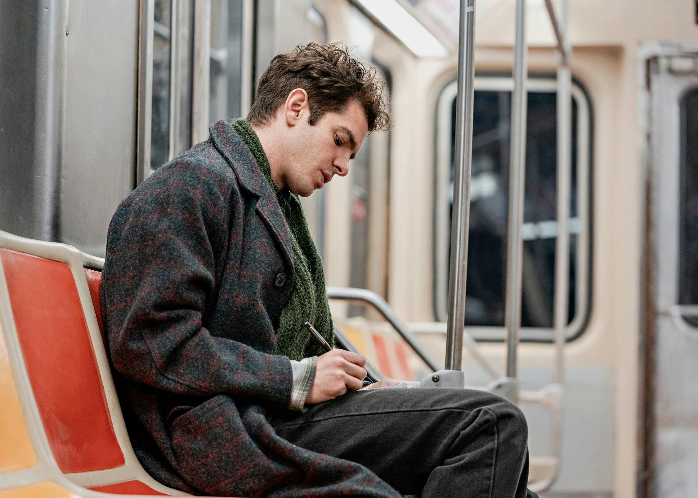 Andrew Garfield as Jonathan Larson writes in his notebook whilst riding the subway.  He wears a grey and red checked long woollen coat, green scarf, and jeans