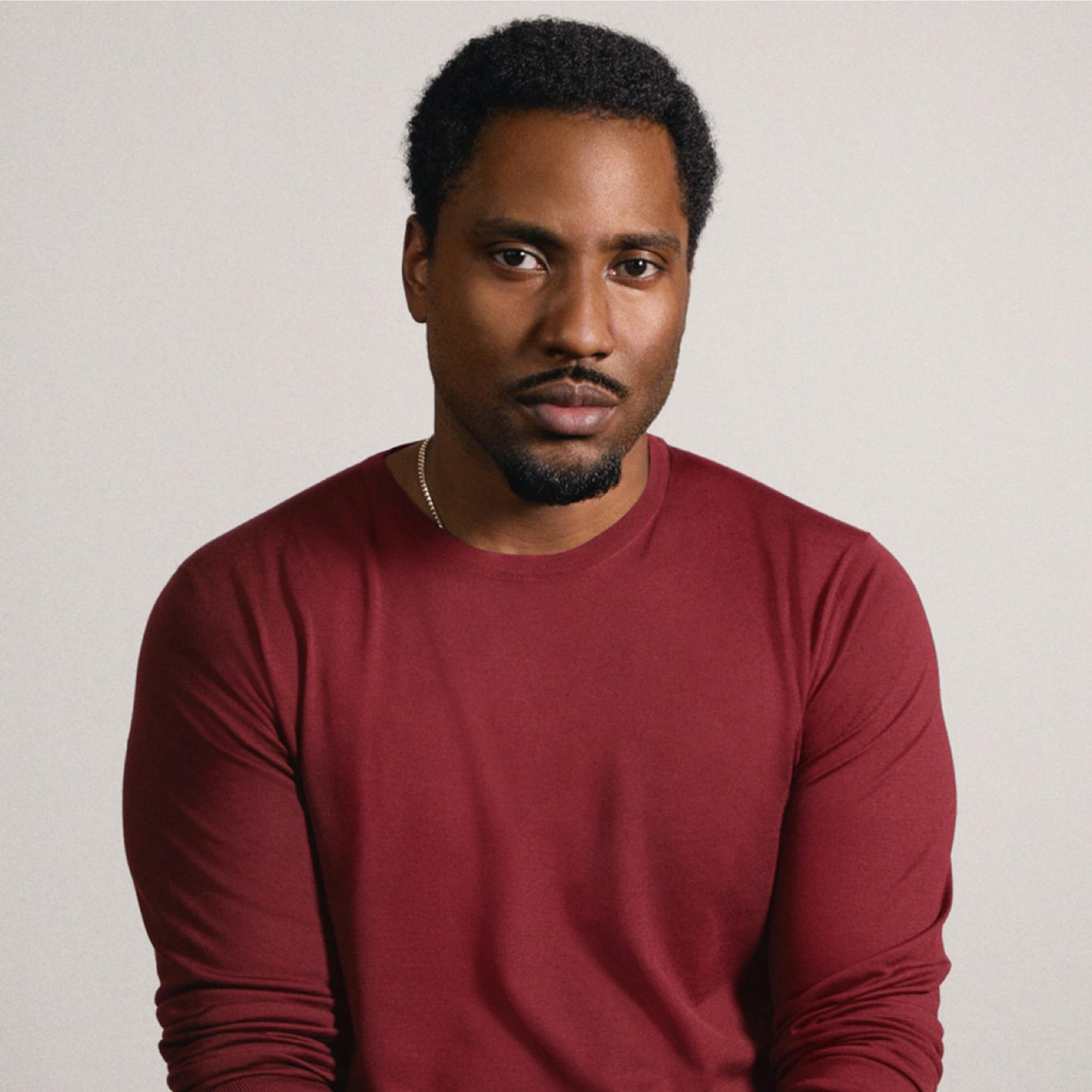 A portrait of John David Washington in a red sweater, set against a beige background. He looks directly to camera.