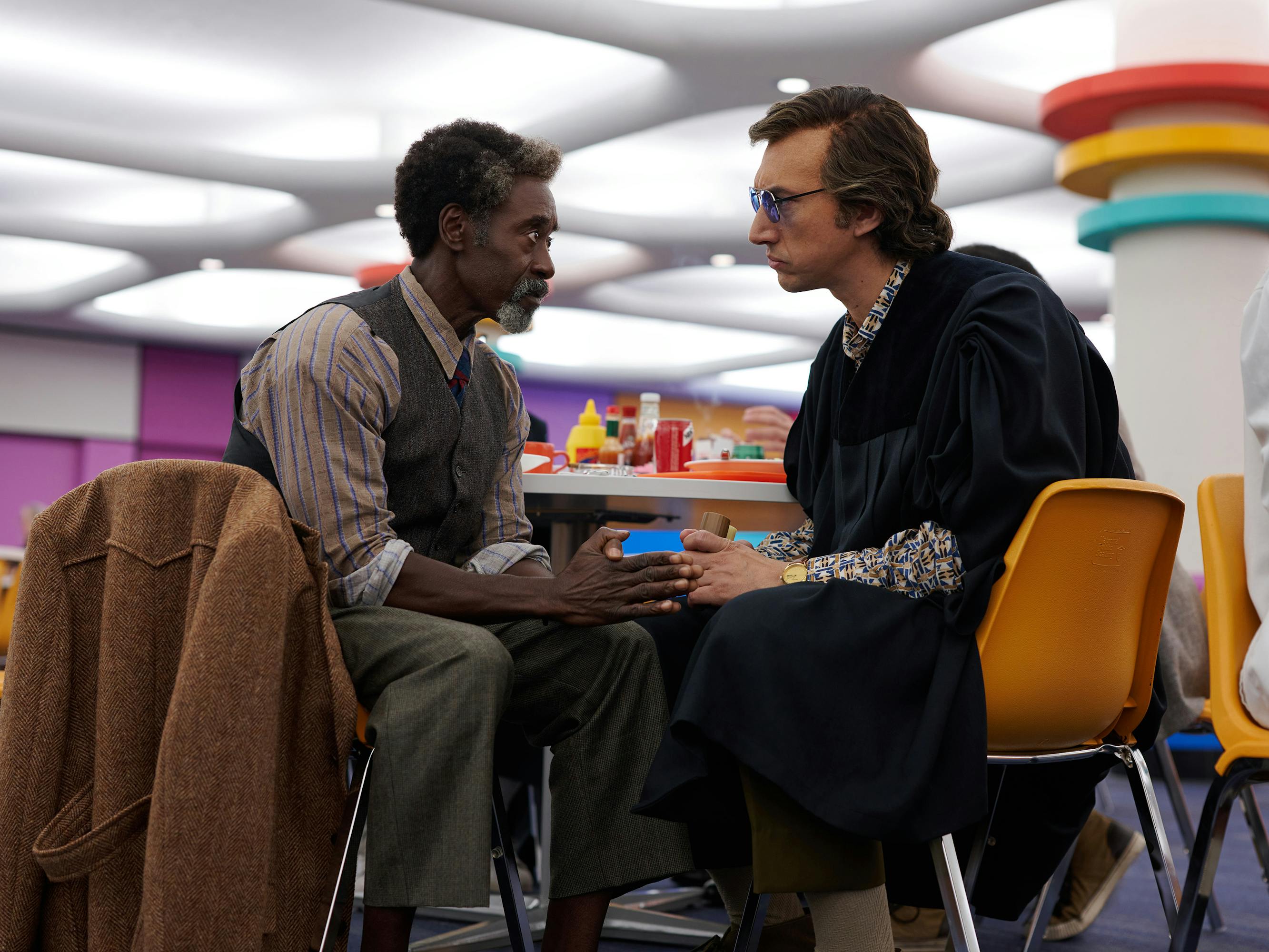 Murray Siskind (Don Cheadle) and Jack Gladney (Adam Driver) sit in yellow chairs at a table in a primary-colored decorated room.