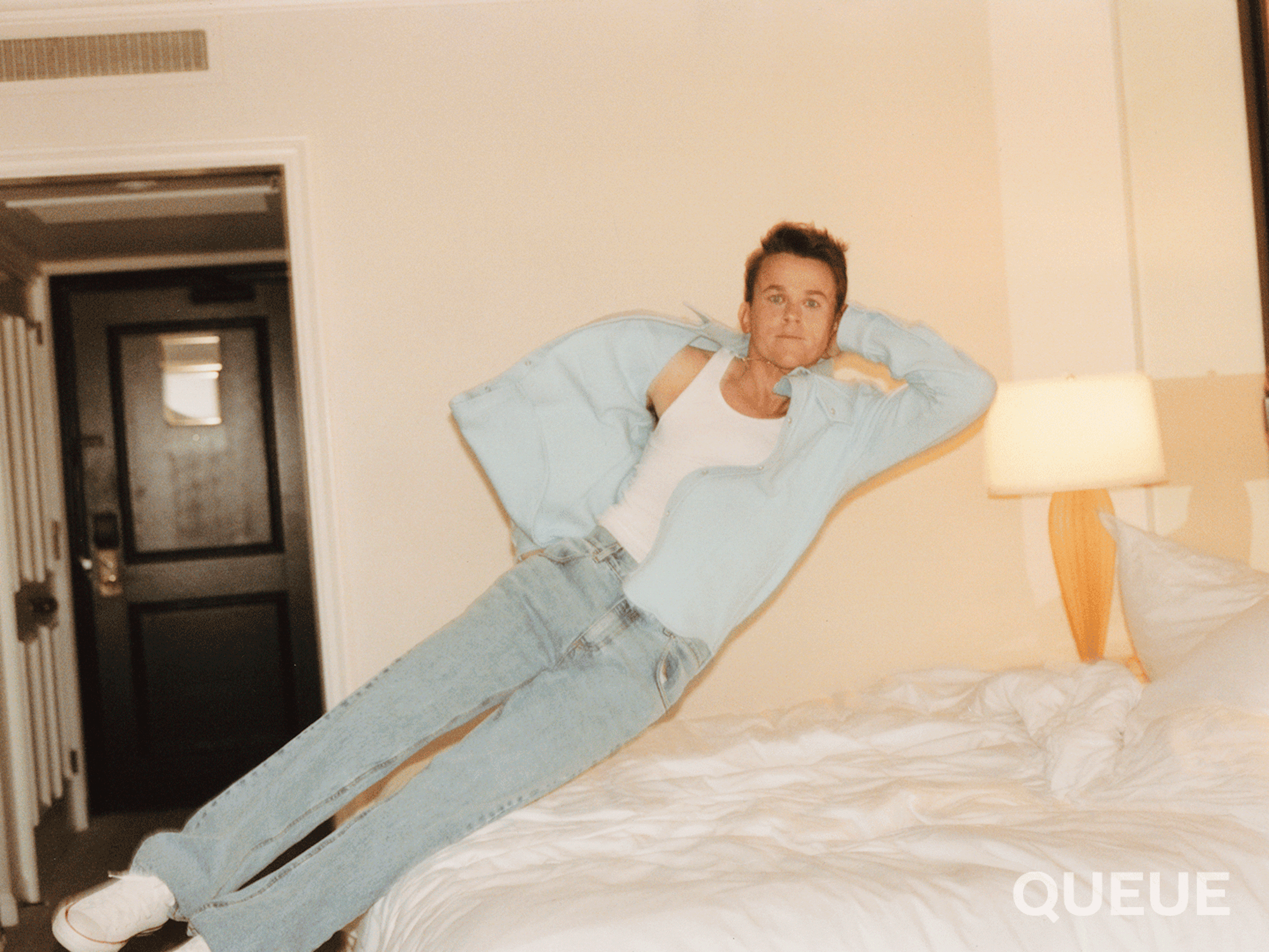 John Owen Lowe flops onto a hotel bed in this short gif. He wears light-wash jeans and a long-sleeved light blue denim shirt over a white tank top. 