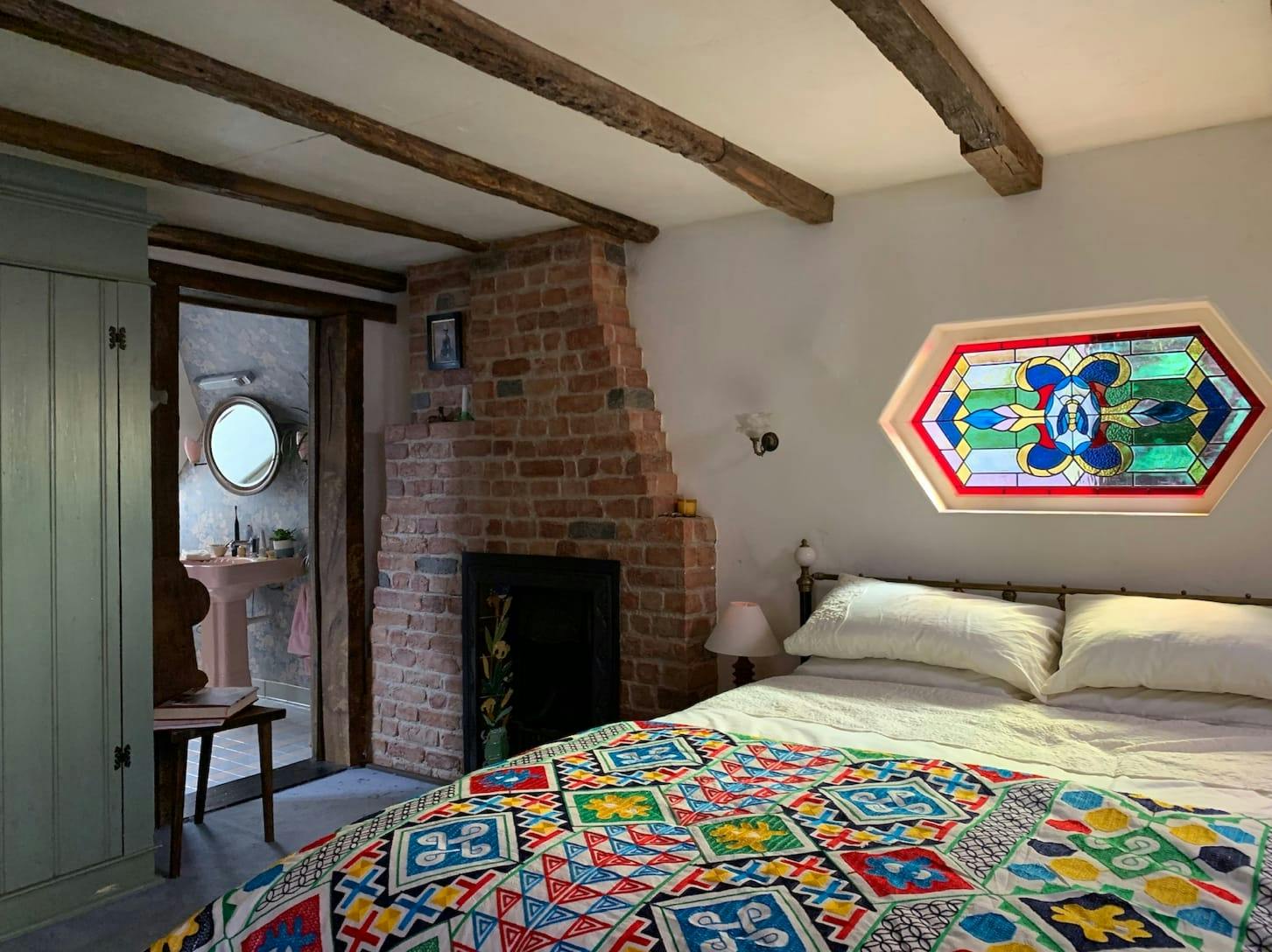 A shot of the bedroom. The bed is predominately white with a multi colored spread, and a multi colored stained glass window. There is a brick fireplace and wood beams on the ceiling.