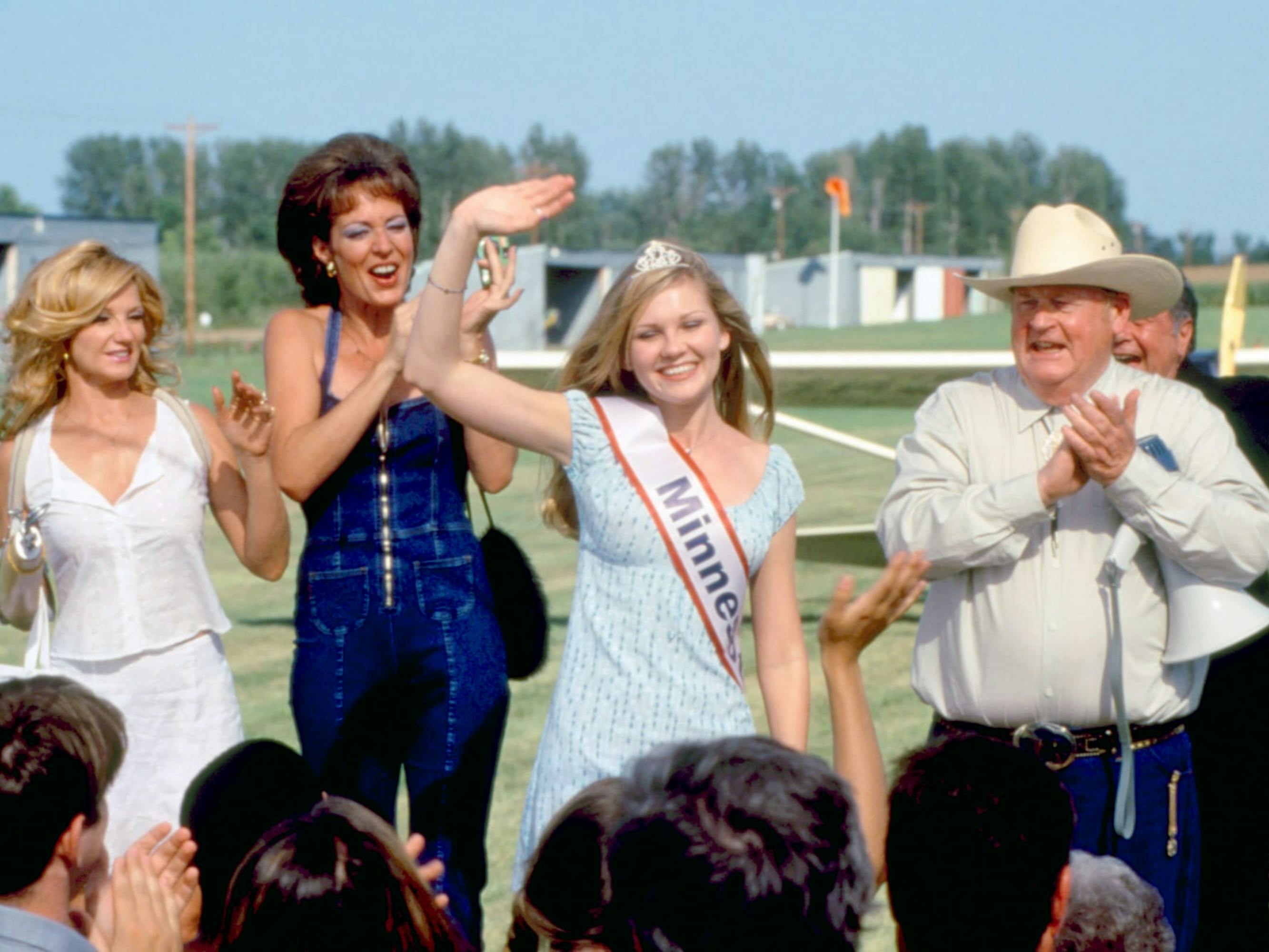 Annette Atkins (Ellen Barkin), Loretta (Allison Janney), and Amber Atkins (Kirsten Dunst) stand together at a beauty pageant. Dunst wears a Minnesota sash and waves at the crowd.