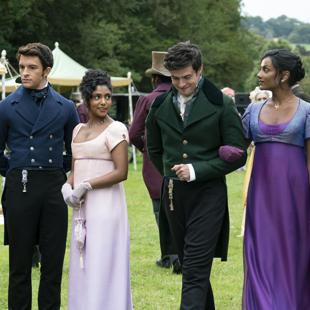 Ruth Gemmell, Jonathan Bailey, Charithra Chandran, Rupert Young, and Simone Ashley stand together outside. Ruth wears a light pink dress, Bailey wears a beautiful navy suit, and Chandran wears a white dress, Young wears a green jacket, and Ashley wears a purple dress.