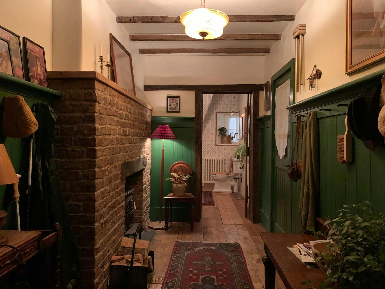 The interior of the house. The walls are finished with green, there is a fireplace, a rug, an overhead light, artwork hanging from the wals, plants, and firewood. The jumbled aesthetic is mostly made up of green and red, and the light makes it very cozy.