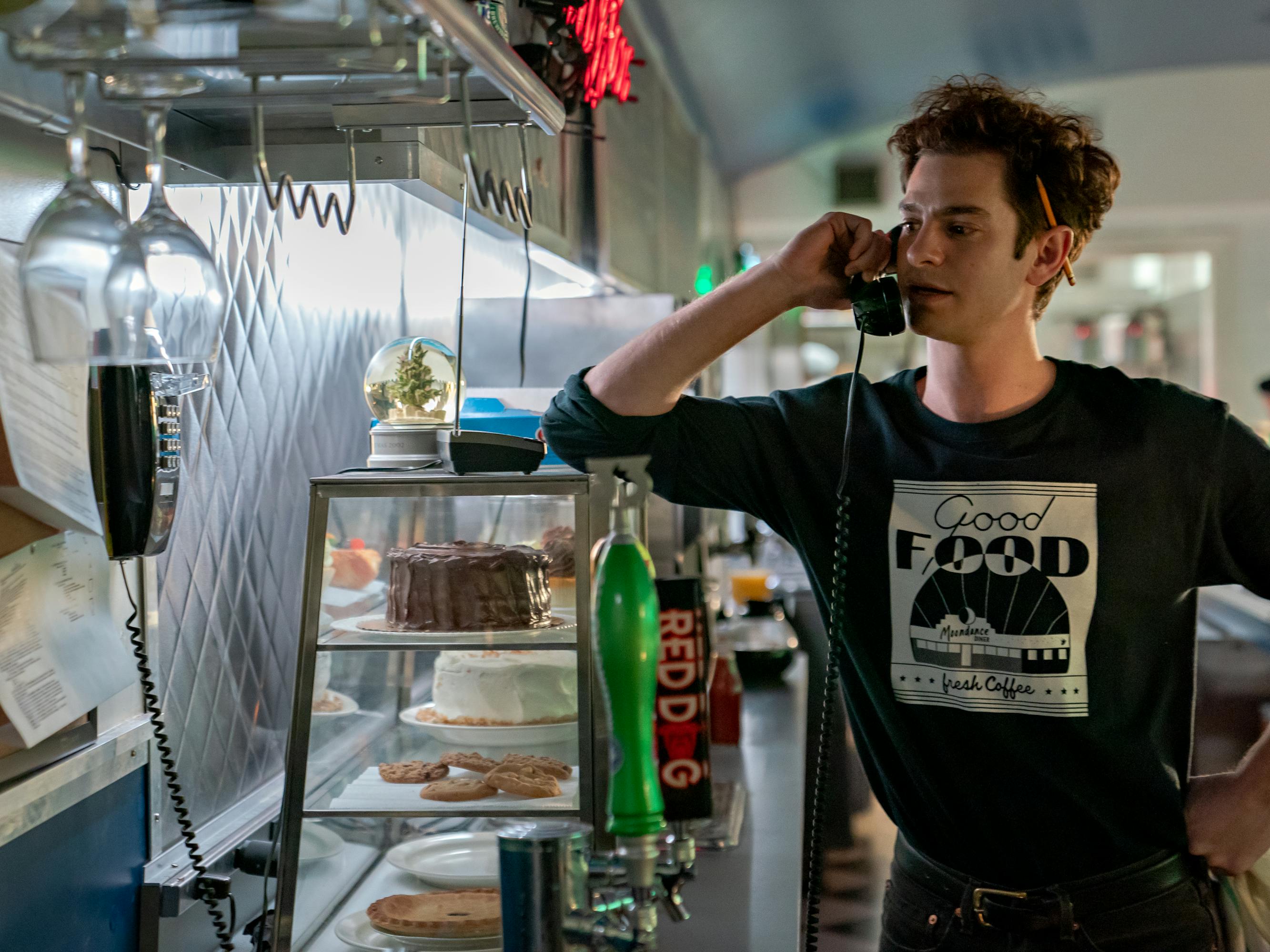 Andrew Garfield wears a Moondance Diner shirt and talks on the phone in the diner. 