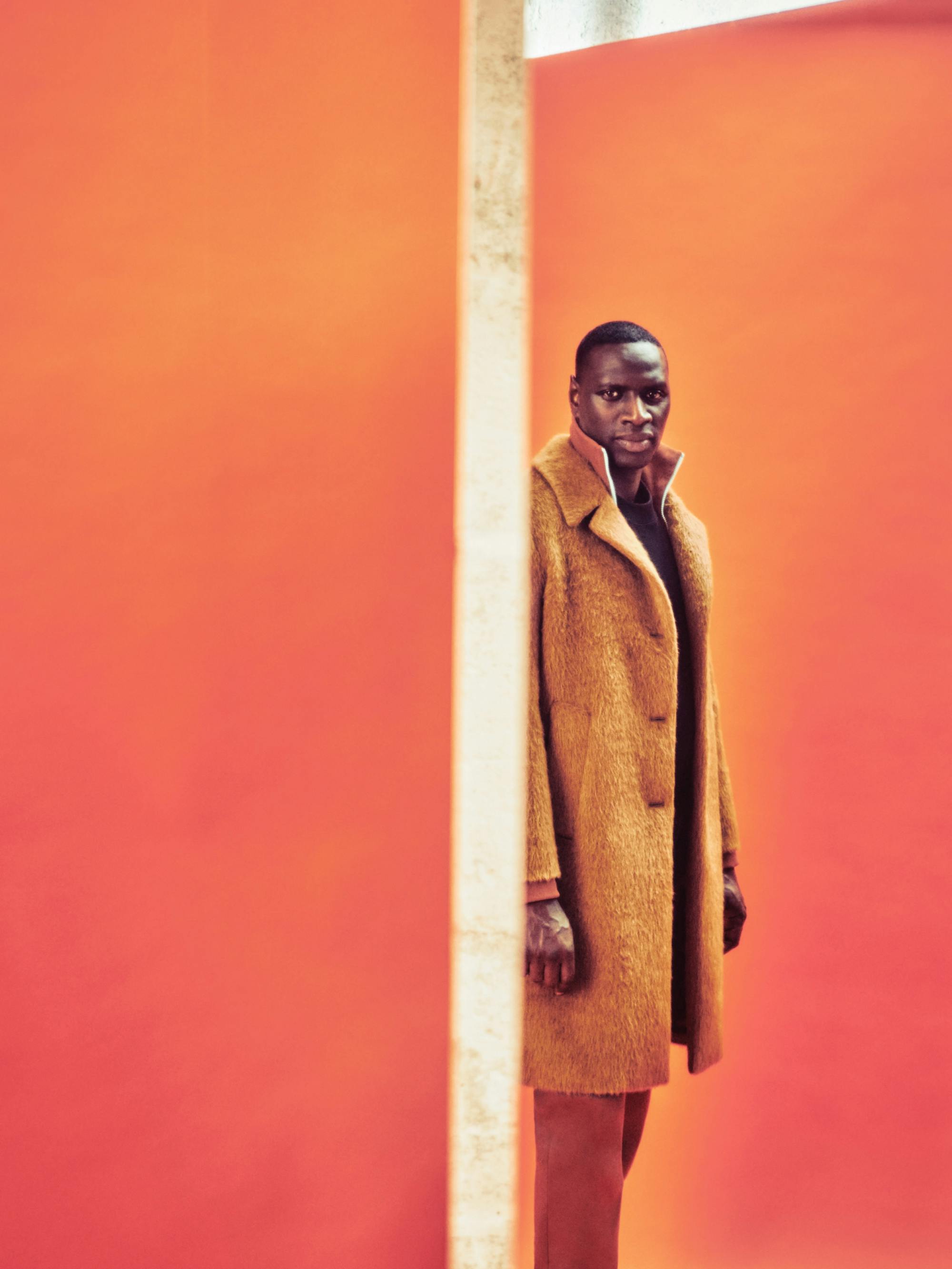 Sy, in a long orange-brown coat, turns to camera.