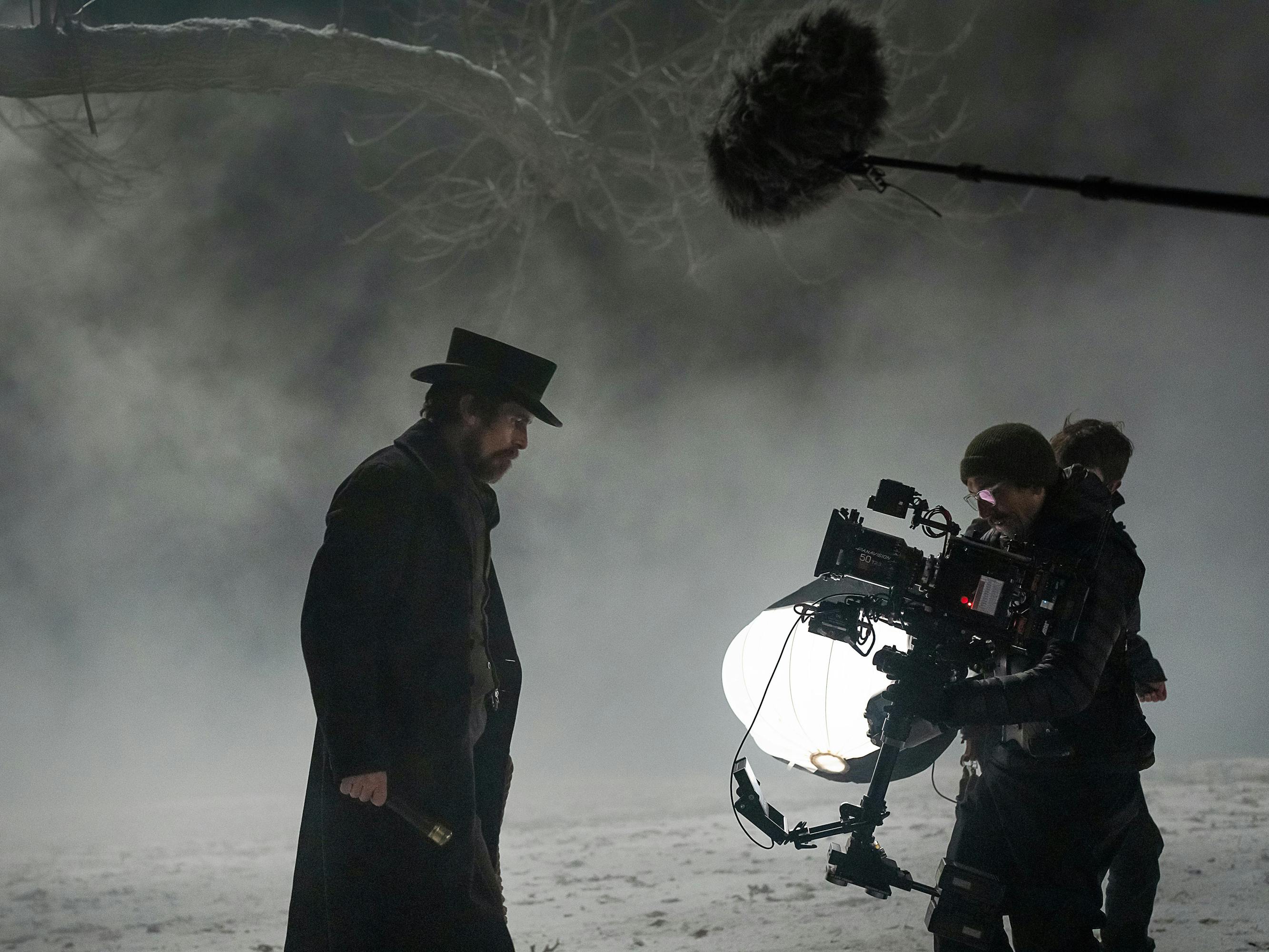 Christian Bale wears a top hat and a dark coat on the set of The Pale Blue Eye. Some crew members stand to his right. The scene is snowy and spooky.