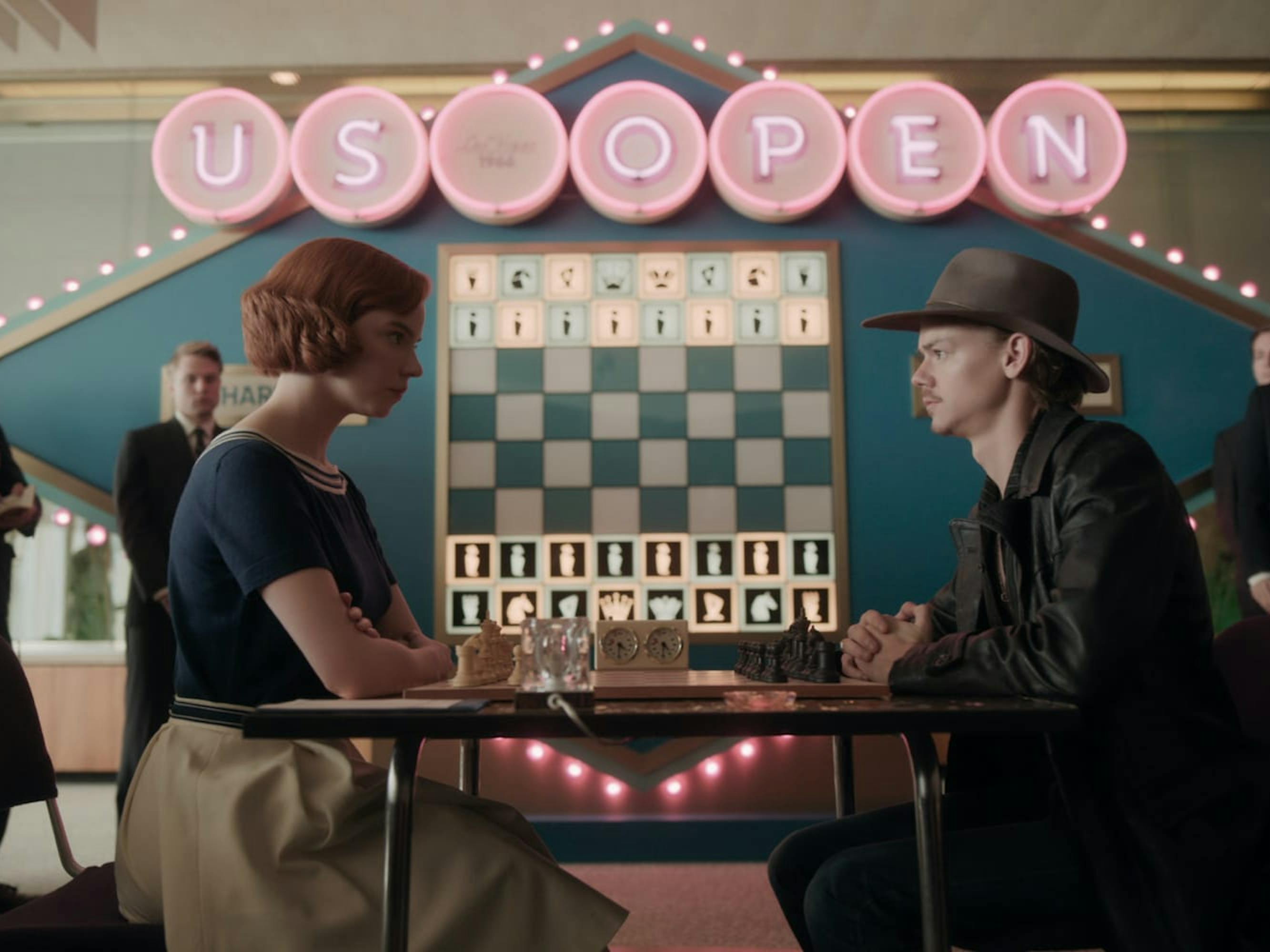 Beth Harmon (Anya Taylor-Joy) and Benny Watts (Thomas Brodie-Sangster) face off in a chess match. Above them reads “US Open” in neon pink light up lettering.