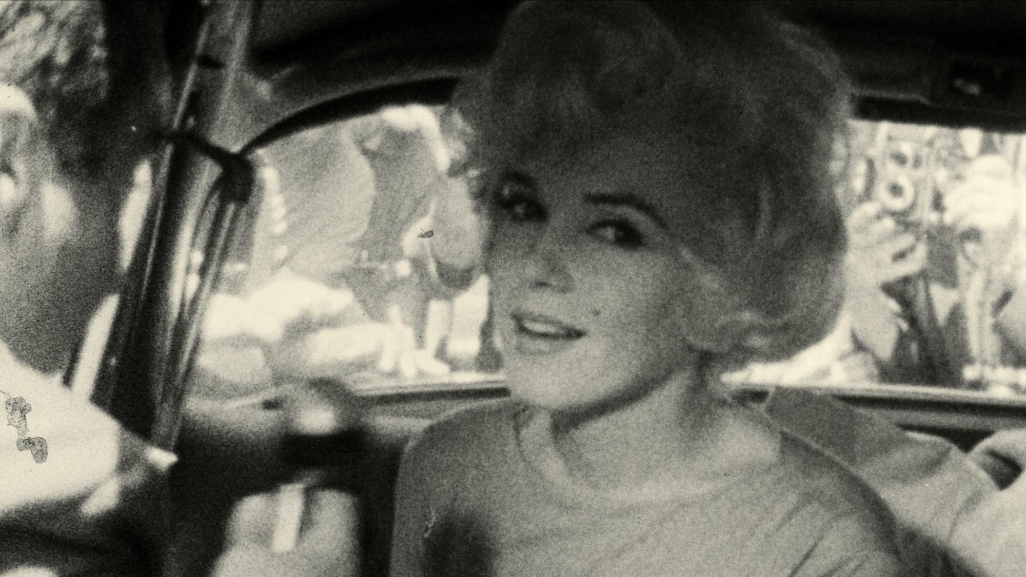 Marilyn Monroe wears a grey top in the back of a car. Through the windshield you can see crowds of people smushed together taking pictures.