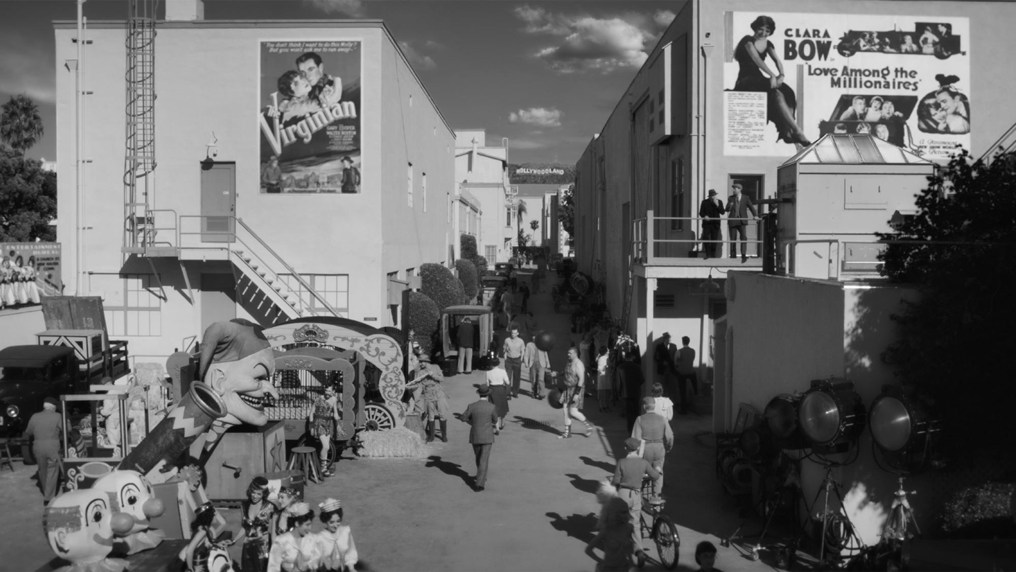 1930s Paramount Studios, recreated for Mank. We look down the gap between two rows of buildings. People wander and mingle, some in costume. A poster for the 1929 Western film The Virginian is pasted to one building. A poster for Love Among the Millionaires starring Clara Bow adorns another. 