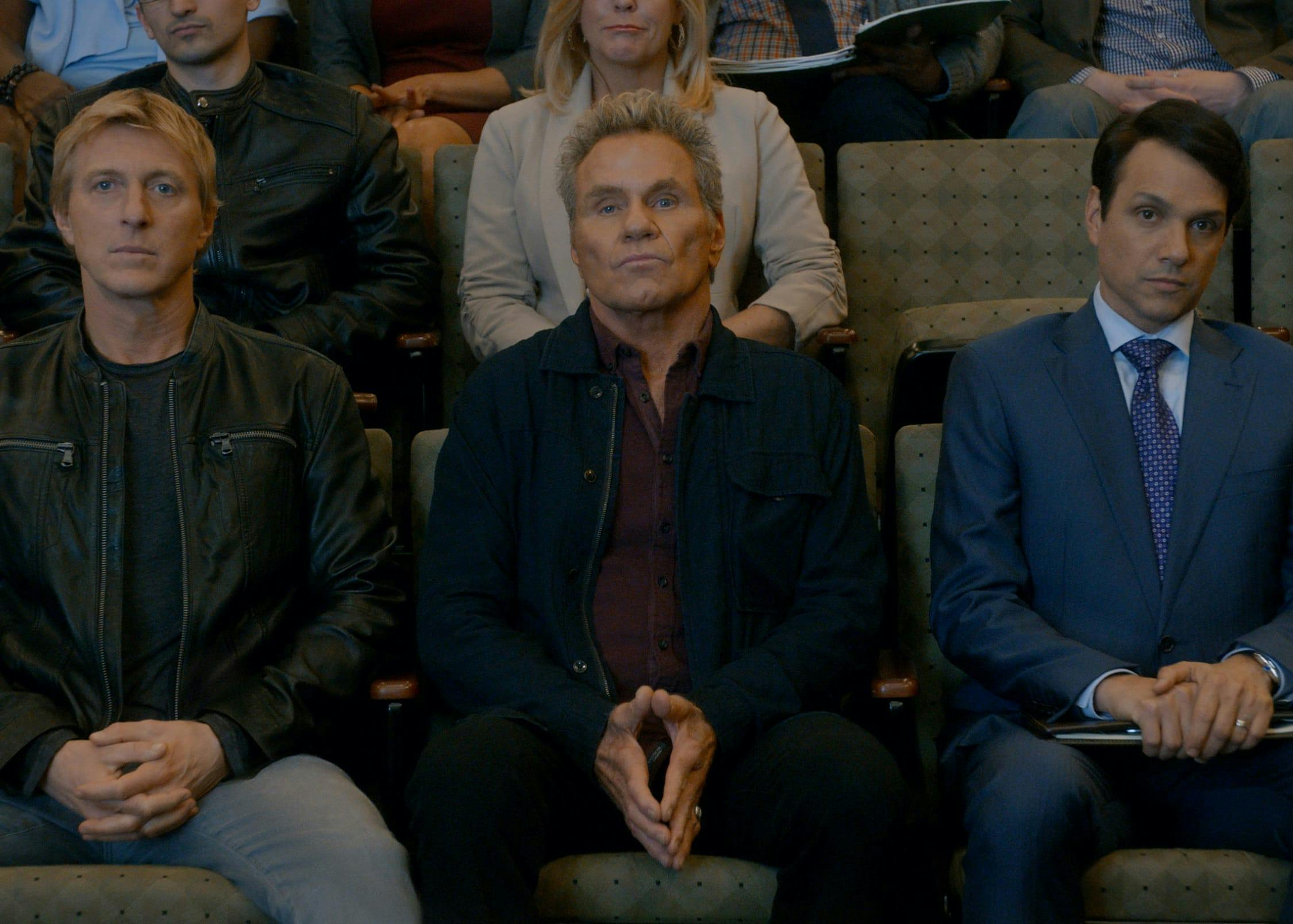 Johnny Lawrence (William Zabka), John Kreese (Martin Kove), and Daniel Larusso (Ralph Macchio) sit in chairs next to each other.