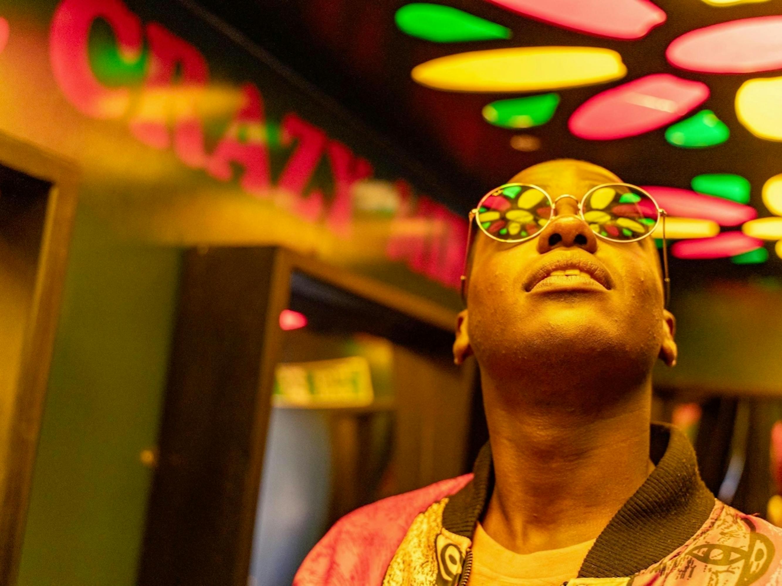 Eric Effiong (Ncuti Gatwa) wears round glasses that reflect the colorful lights that are being projected onto the ceiling. In the background reads “Crazy” in red font.