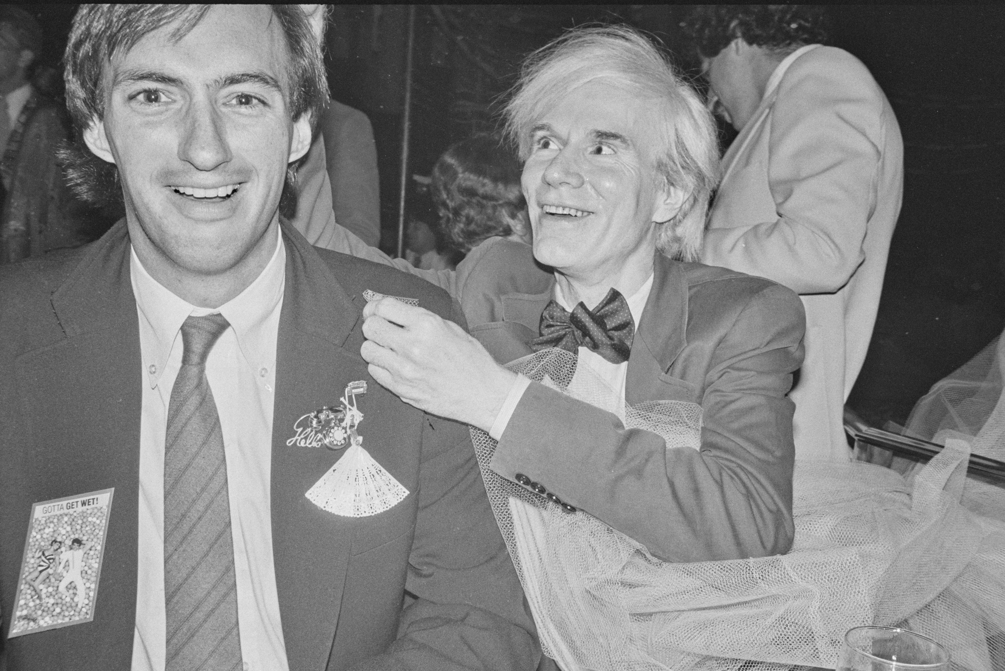 Jon Gould and Andy Warhol sit together. Jon wears a dark blazer with many eccentric pins. Warhol grins and looks at something off camera. He wears a bowtie and a sits in a pool of tulle.