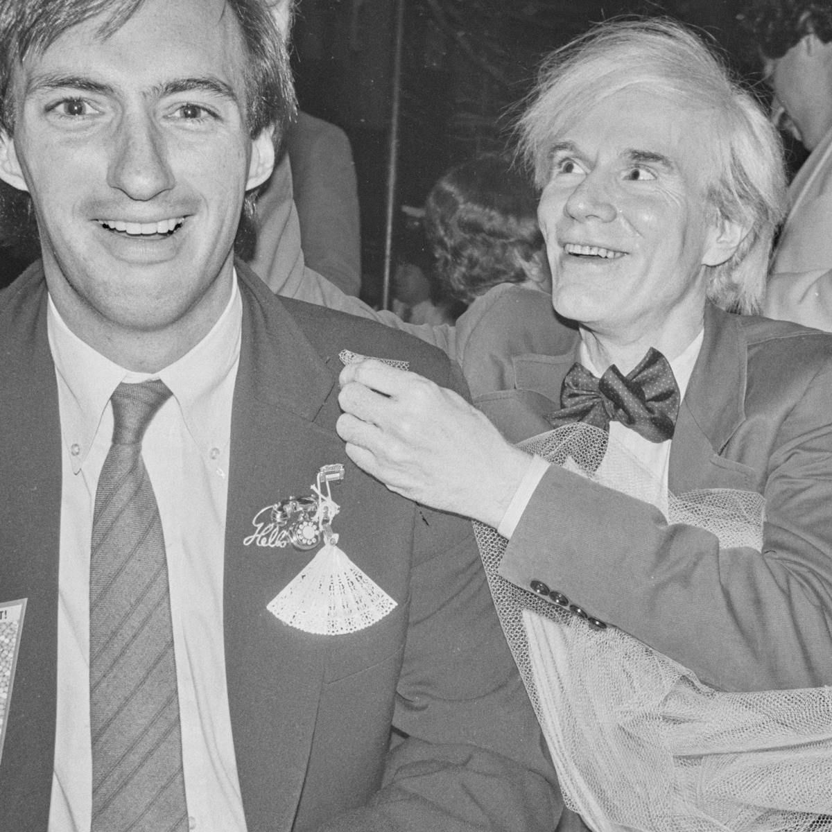 Jon Gould and Andy Warhol sit together. Jon wears a dark blazer with many eccentric pins. Warhol grins and looks at something off camera. He wears a bowtie and a sits in a pool of tulle.