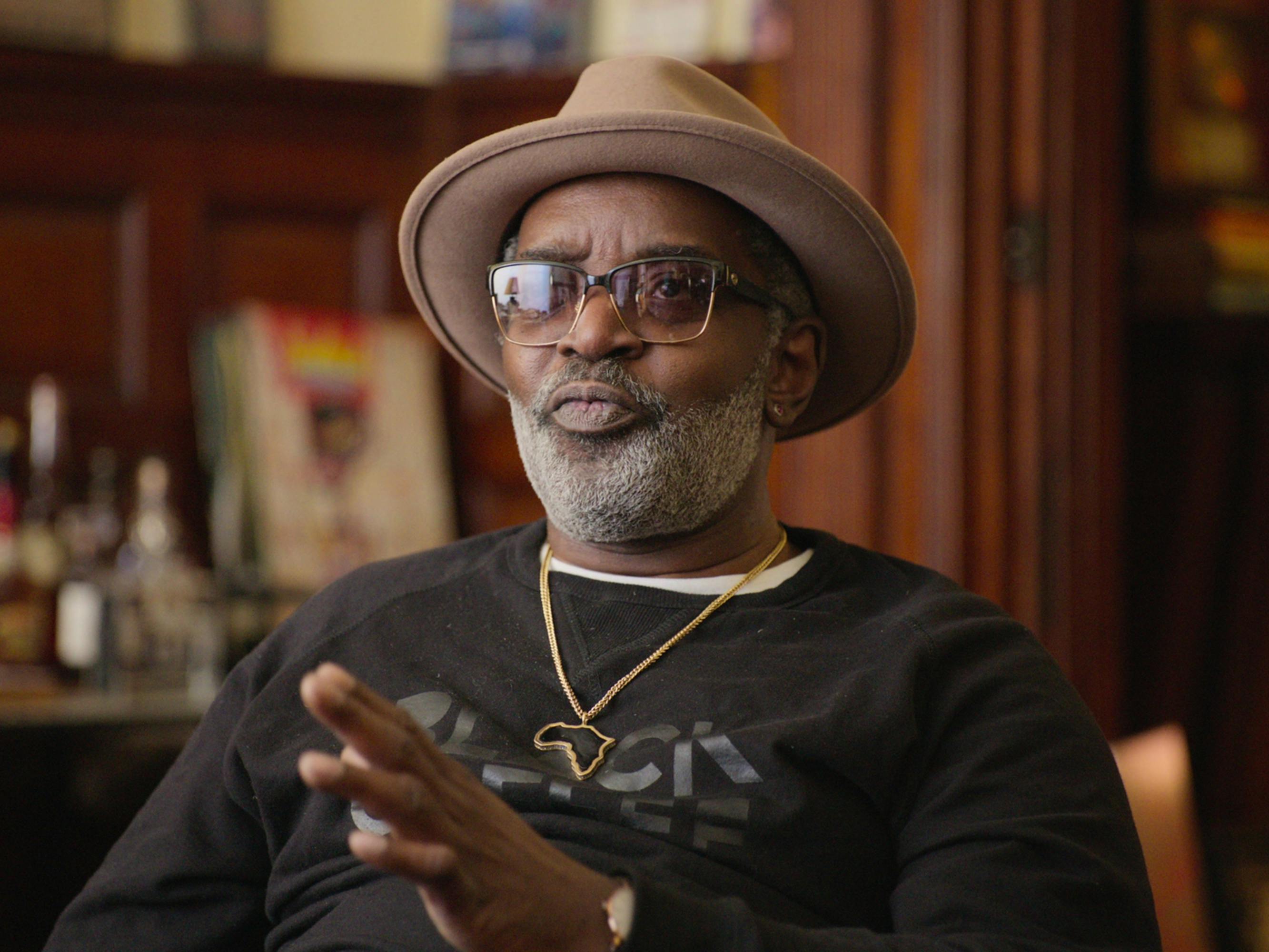 Fab 5 Freddy wears a brown hat and black shirt. He sits in a wood paneled room.