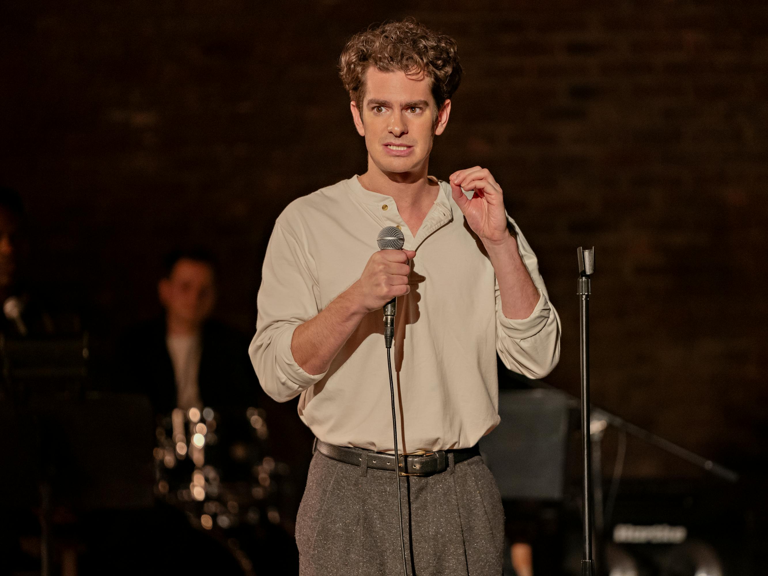 Andrew Garfield wears a white shirt and grey slacks and stands on a darkened stage holding a mic.
