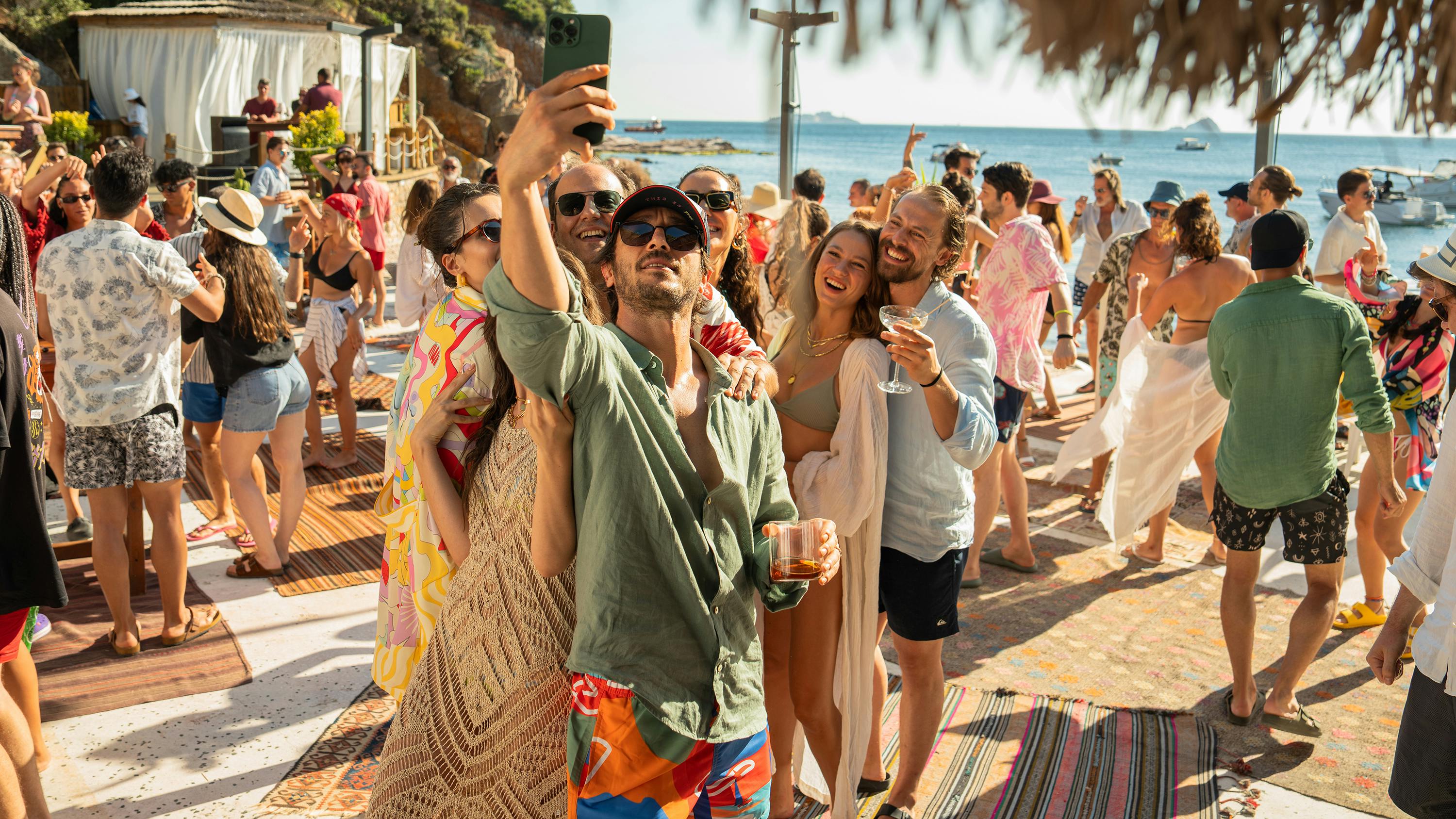 A busy, colorful beach party with the blue sea in the background. A group of friends huddle together to take a selfie. 