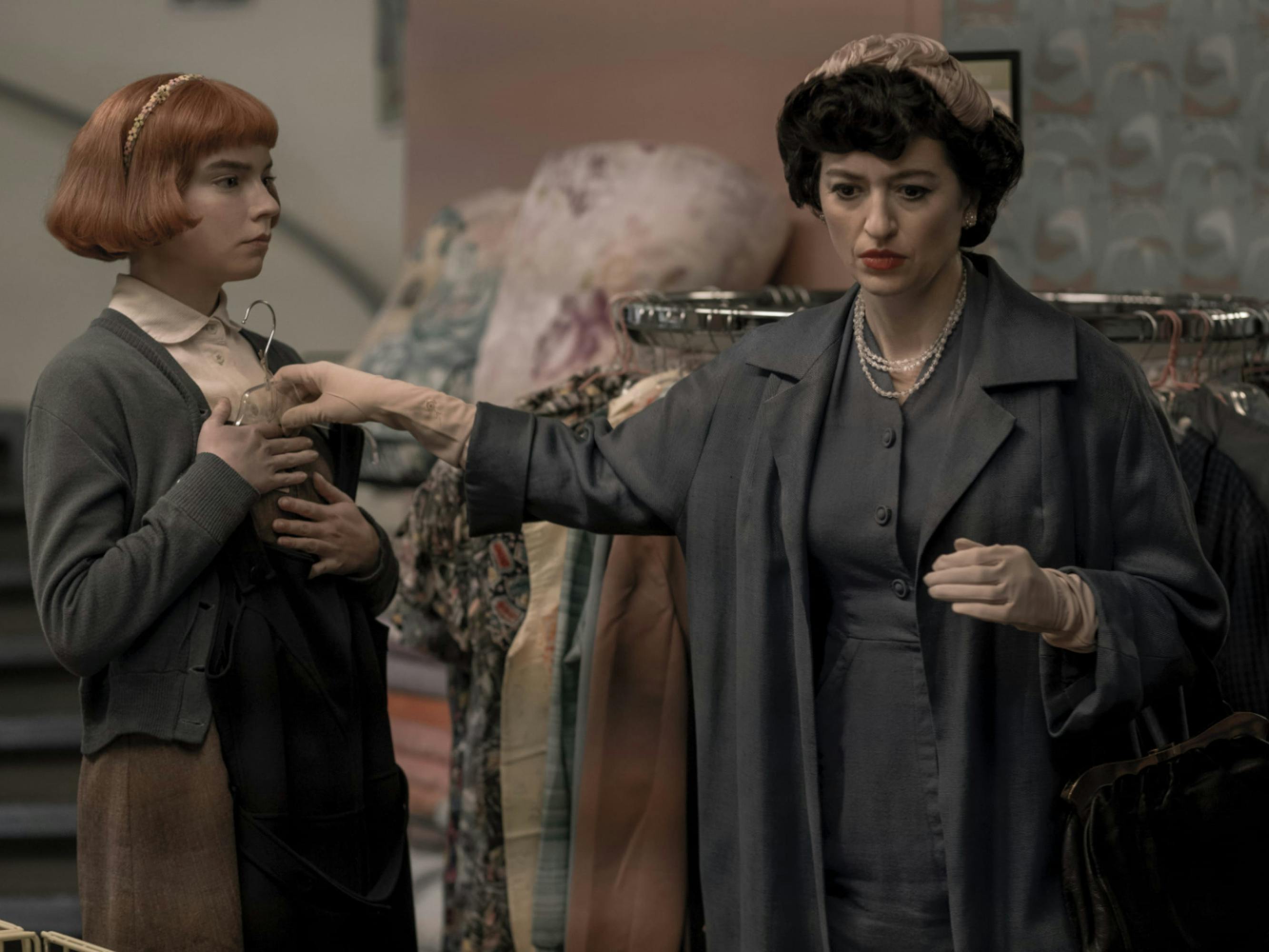 Alma Wheatley, played by Marielle Heller, holds up a dress to her adoptive daughter Beth, played by Anya Taylor-Joy. Beth has a short red bob haircut and is wearing a gray sweater. Alma is in an all gray outfit with a pearl necklace and a pink hair bow. They are standing in front of a clothing rack in a store.