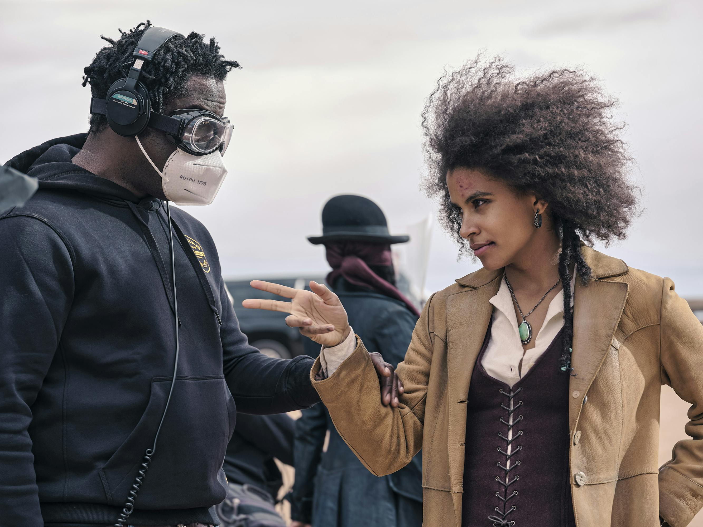 Jeymes Samuel and Zazie Beetz stand together outside. Samuel wears a black sweatshirt, black earphones, googles, and a white N95 mask. Beetz wears a maroon corset, tan jacket, and white shirt.