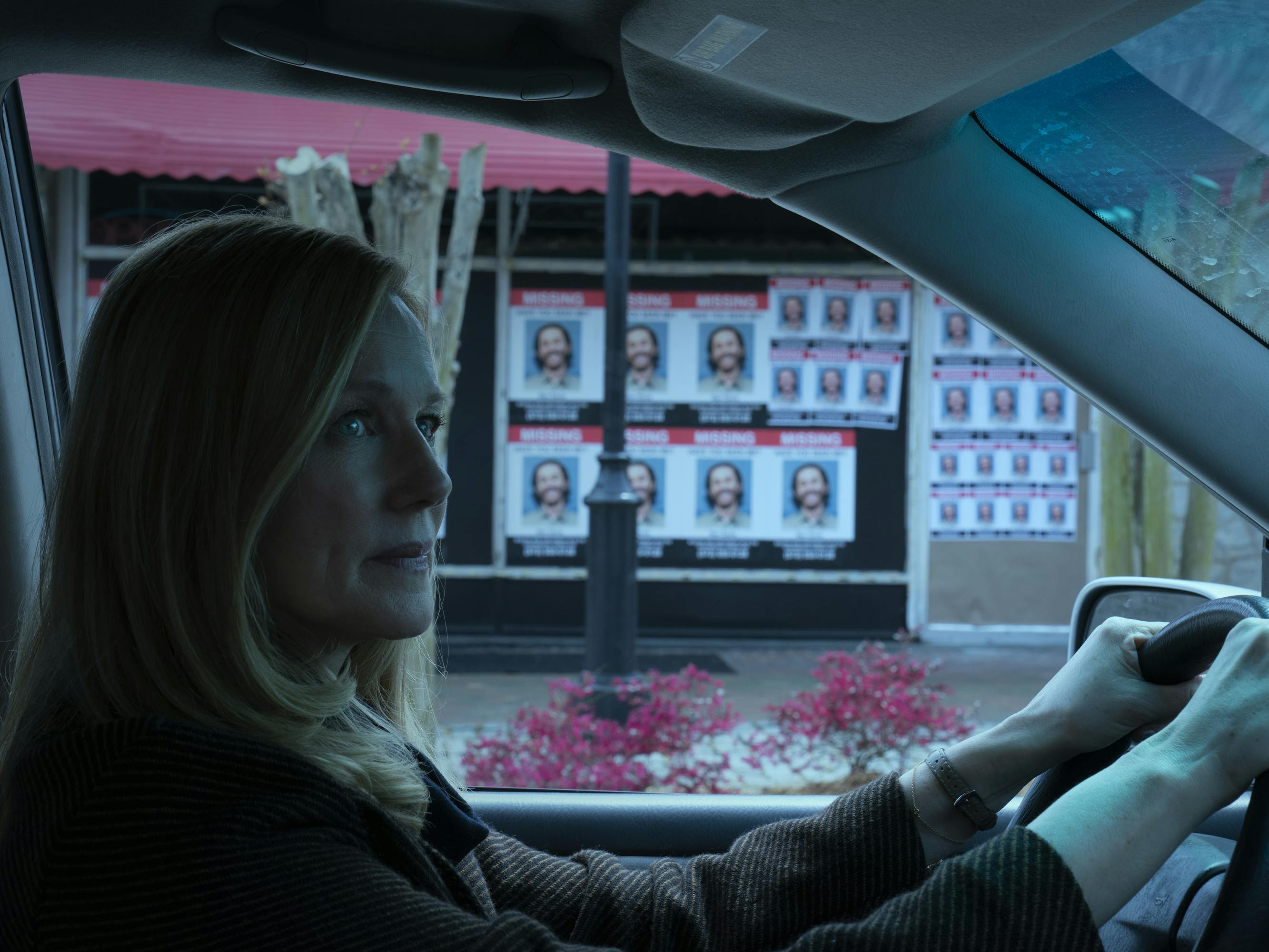Wendy Bryde (Laura Linney) looks nervous as she drives her car past some wanted posters of her brother. The scene is eerie and grey. 