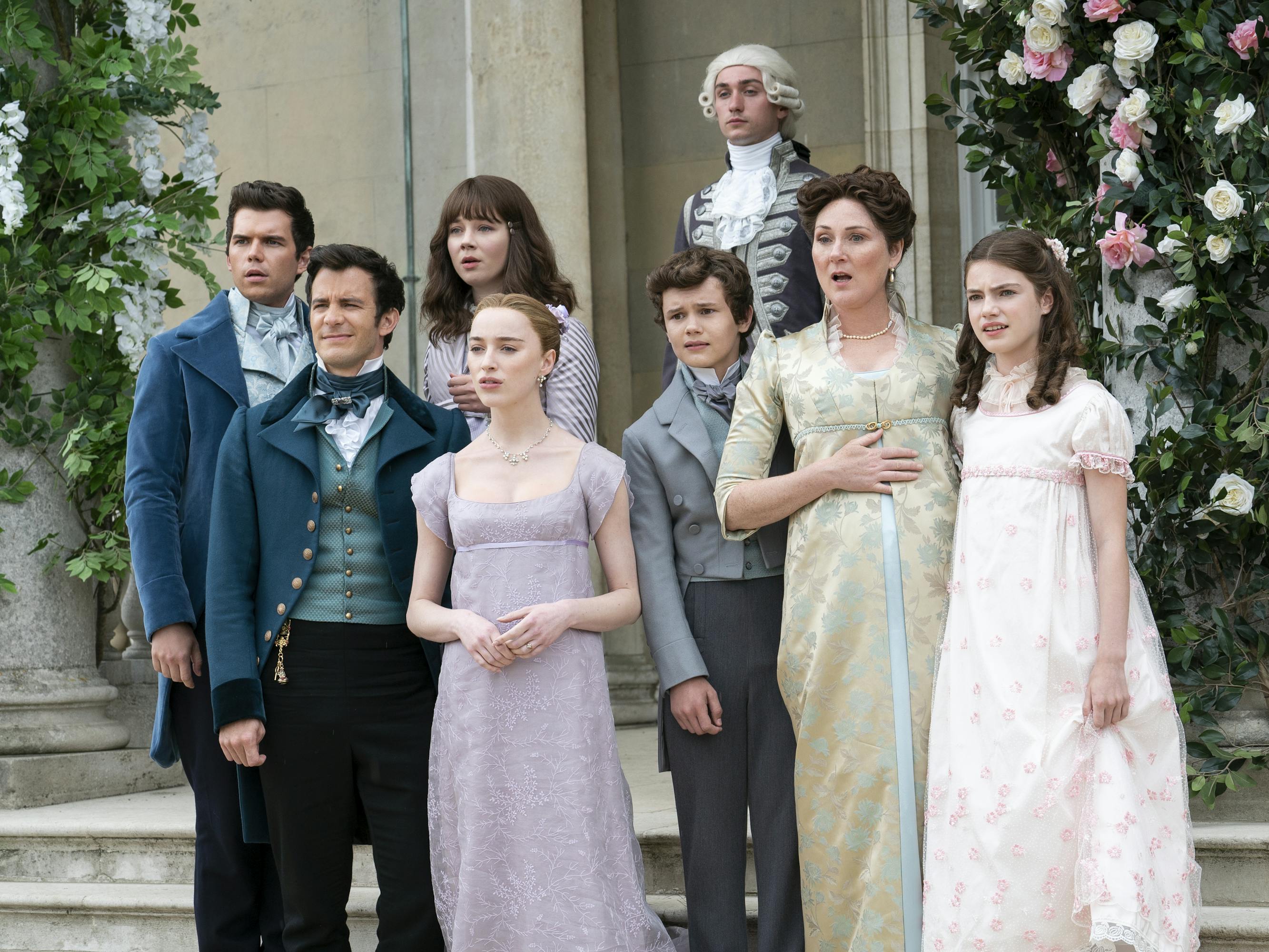 Luke Newton, Luke Thompson, Claudia Jessie, Phoebe Dynevor, Ruth Gemmell, Florence Hunt stand together outisde. The men wear dark blazers with high necked shirts. The women wear dresses in shades of purple, pink, and yellow.